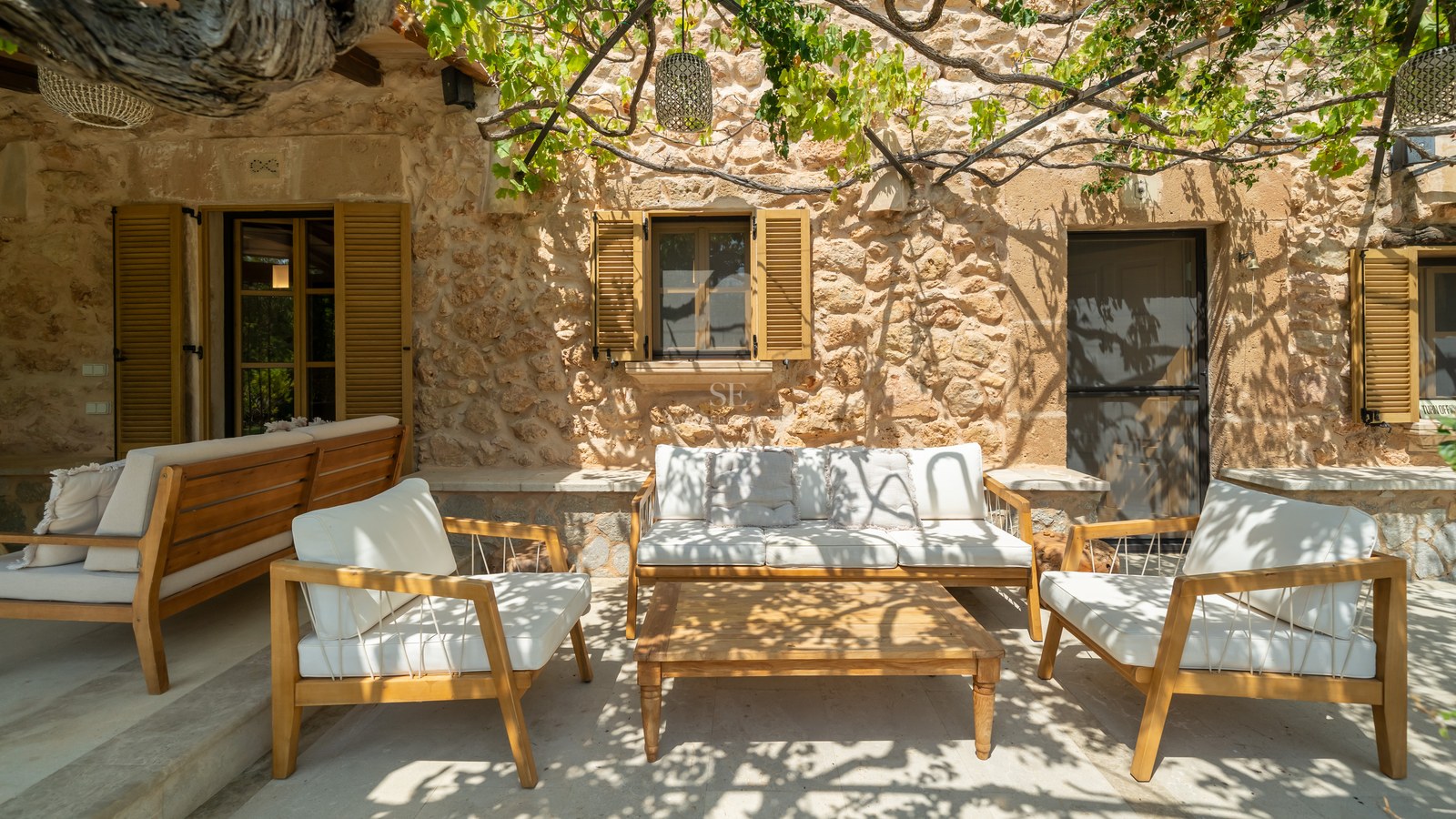 Sun-drenched terrace with wooden lounge furniture, natural stone walls, and a grapevine-covered pergola.