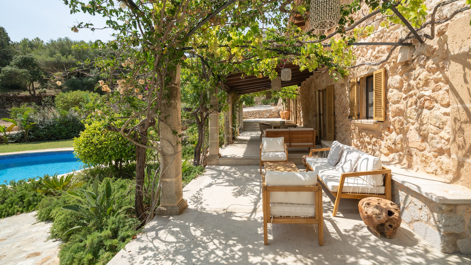 Sun-dappled stone terrace with white cushioned wooden seating, lush vines, and a blue pool in the background.