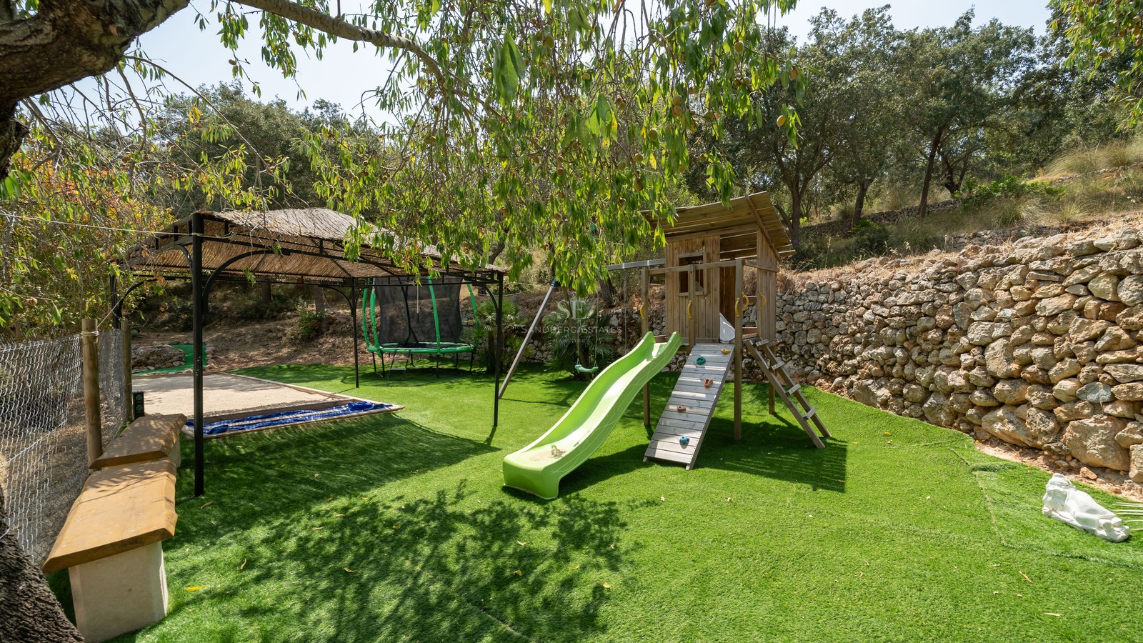 Children's wooden playhouse with a green slide, trampoline, and thatched gazebo on artificial grass bordered by a stone wall.