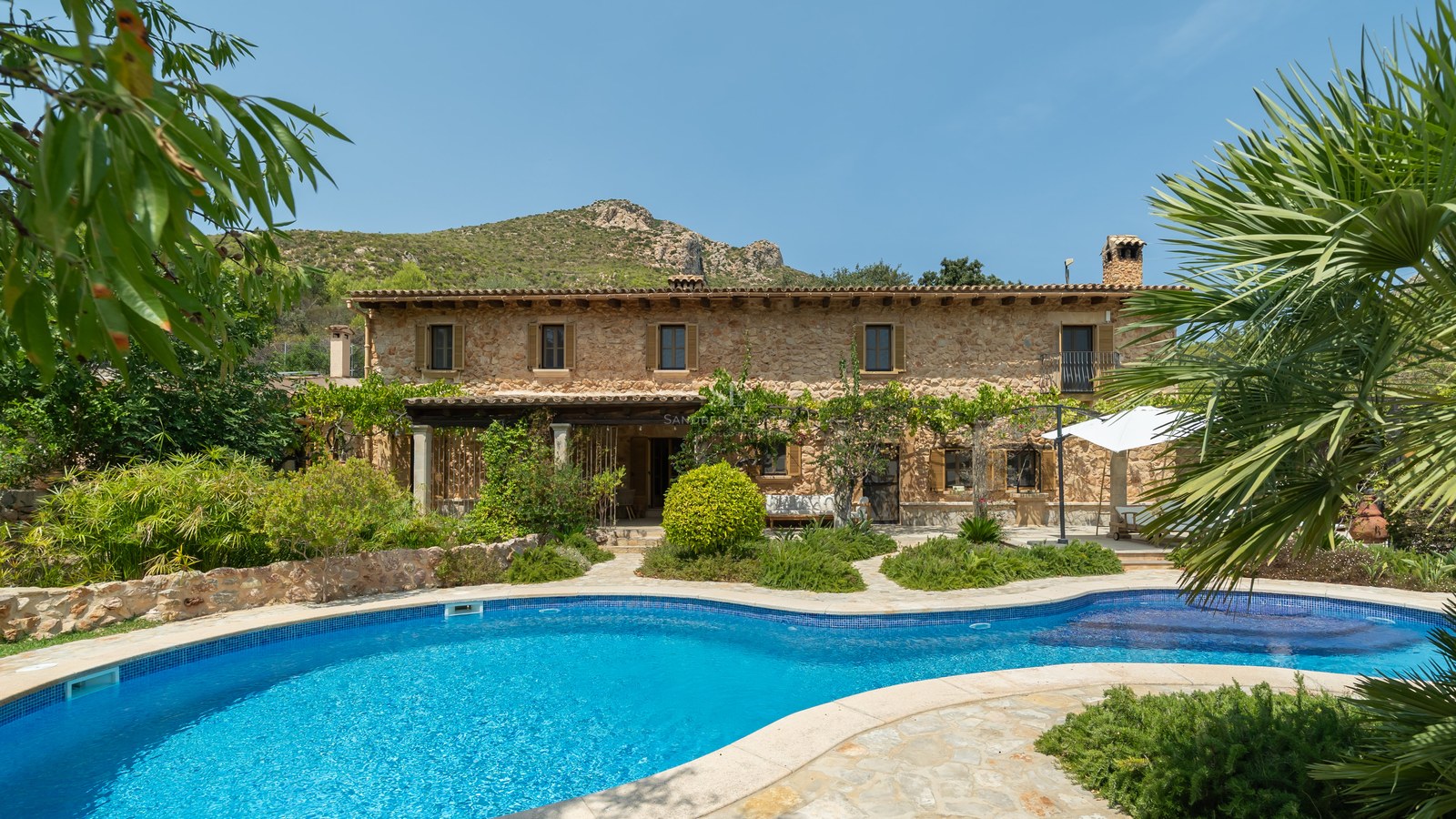 Large natural stone villa with a curved blue swimming pool, lush gardens, and mountain views under a clear sky.