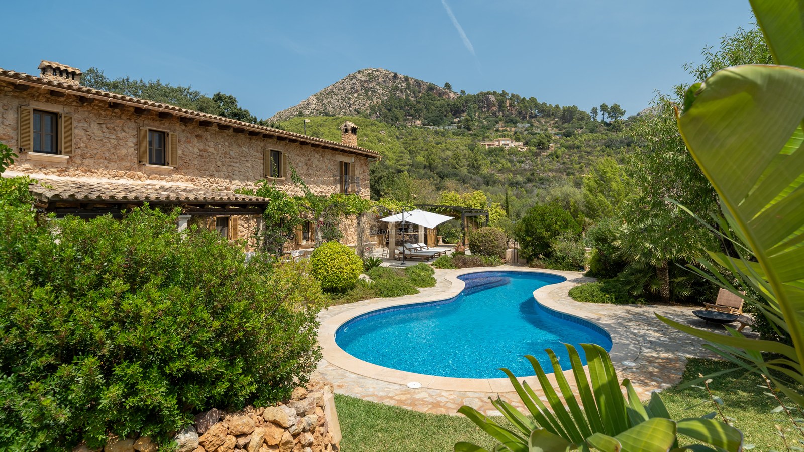 Luxury stone farmhouse with a curved blue swimming pool, lush green gardens, and a scenic mountain backdrop.