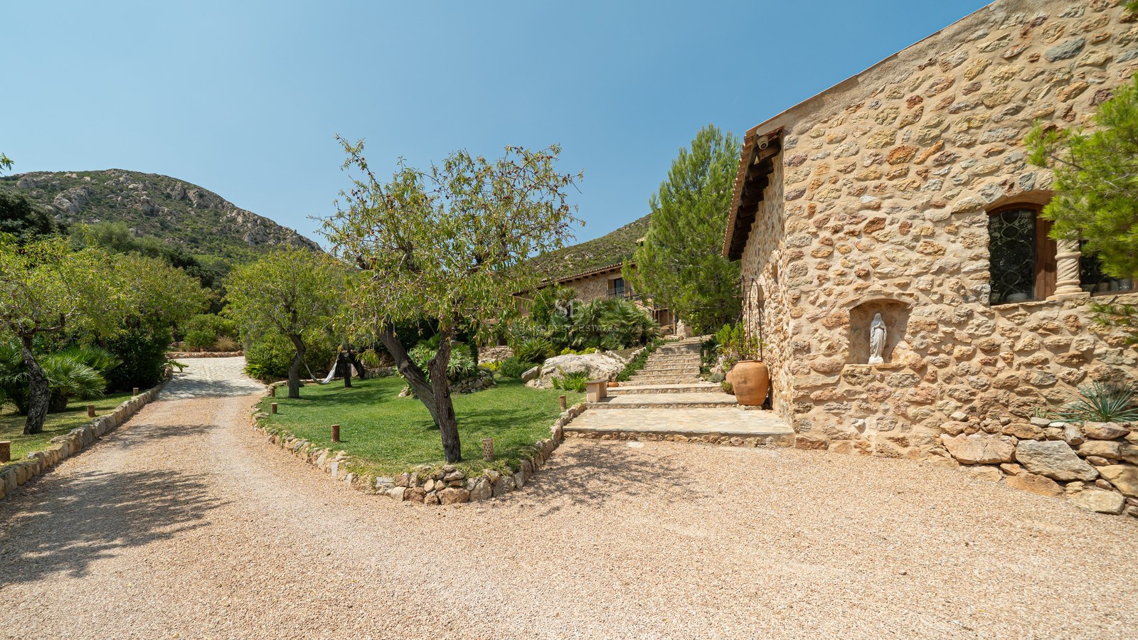Natural stone villa exterior with gravel driveway, manicured gardens, and mountain backdrop under a clear blue sky.