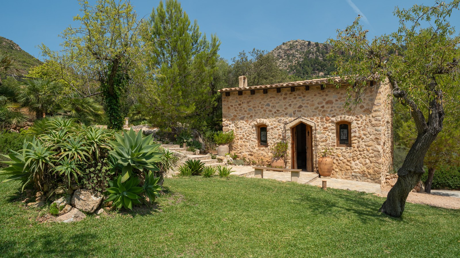Stone cottage with terracotta roof nestled in a Mediterranean garden against a mountain backdrop.