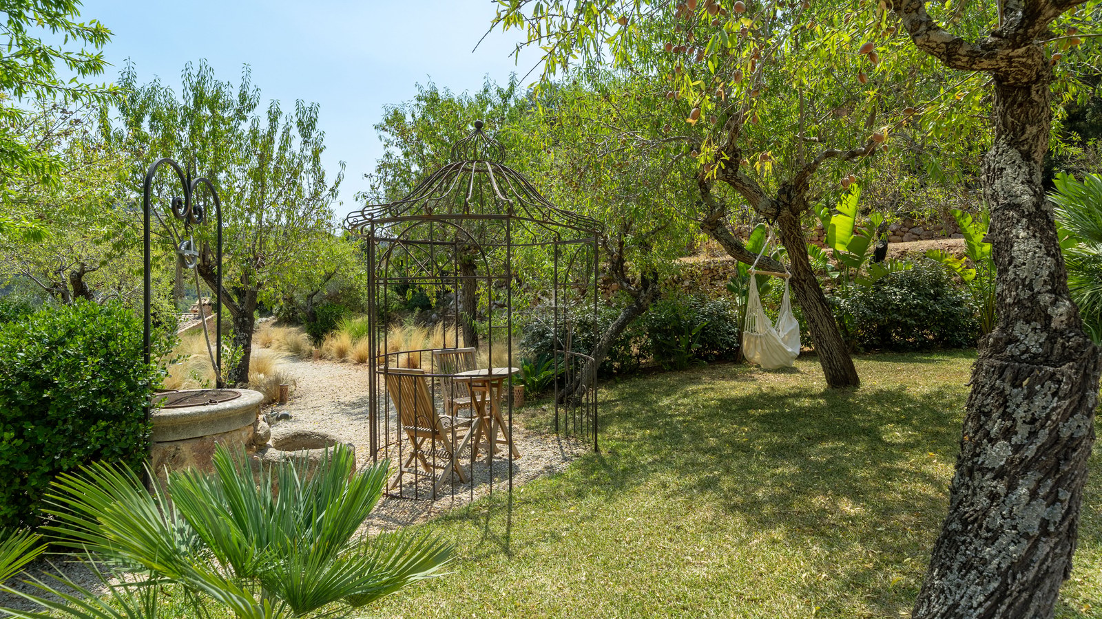 Wrought iron gazebo and stone well in a lush Mediterranean garden with a white hammock hanging from a tree.