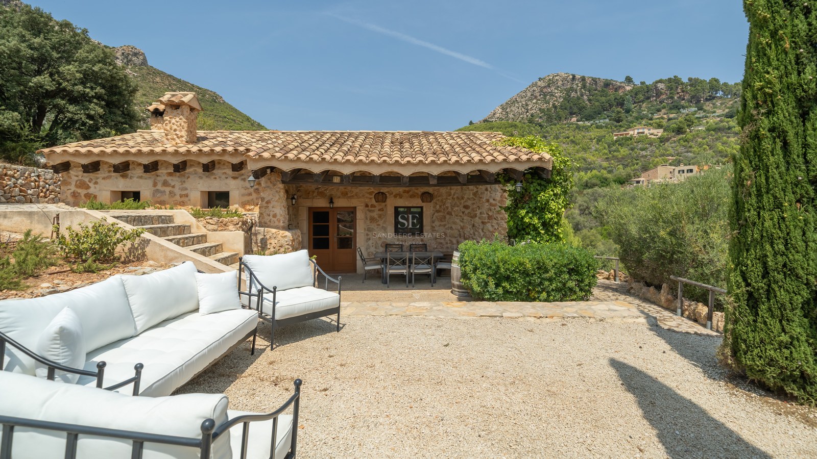 A rustic stone villa with a gravel terrace featuring white sofas and a covered dining area against a mountain backdrop.
