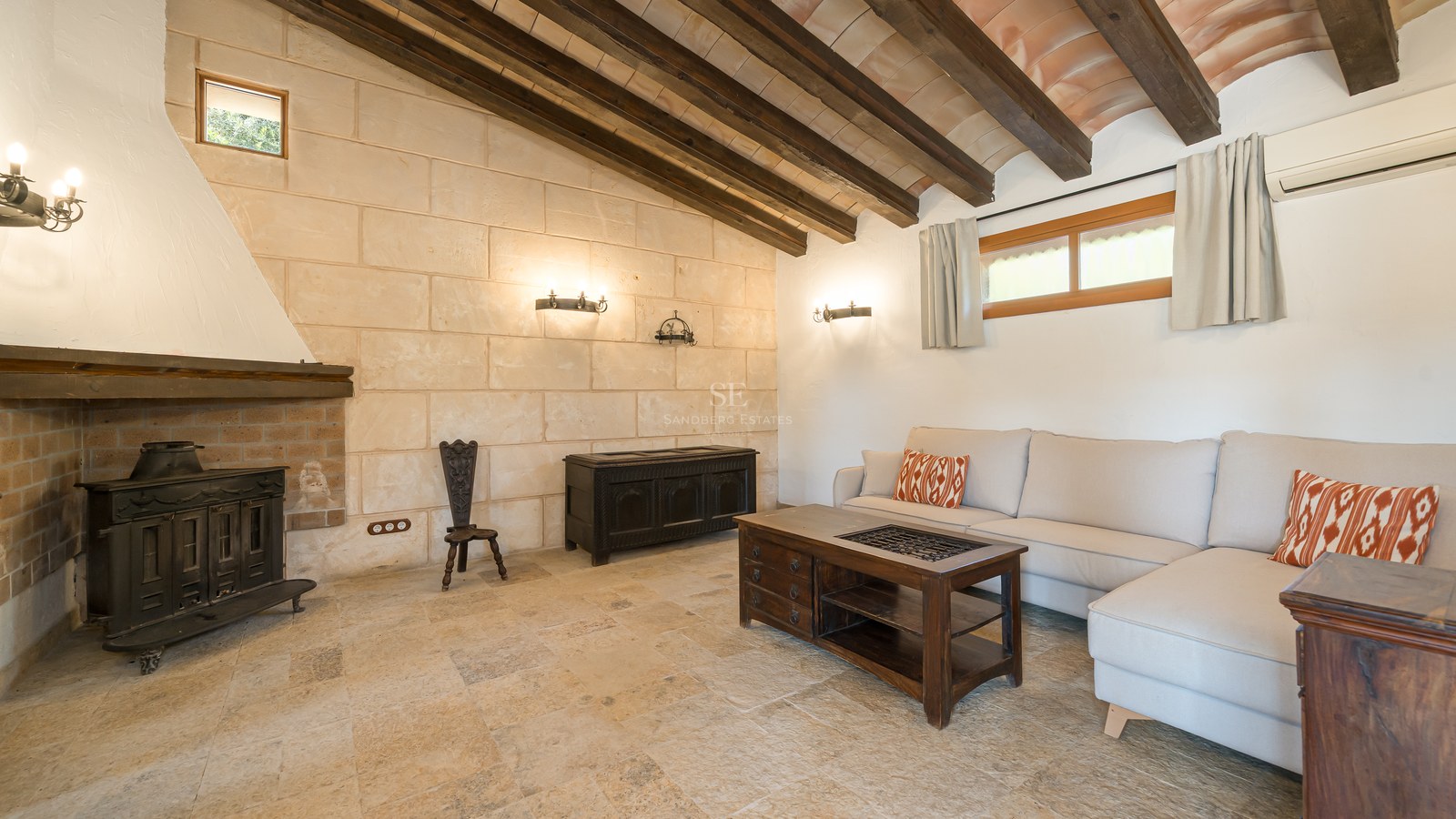 Rustic living room featuring a stone fireplace, exposed wooden ceiling beams, stone floors, and a modern beige sofa.