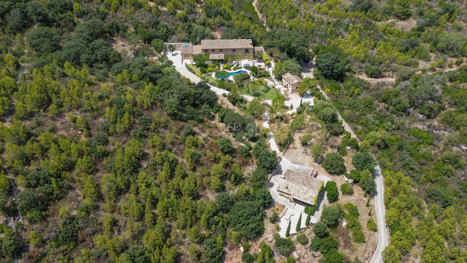Vista aérea de una gran finca de piedra con piscina y casa de invitados rodeada de un denso bosque mediterráneo.