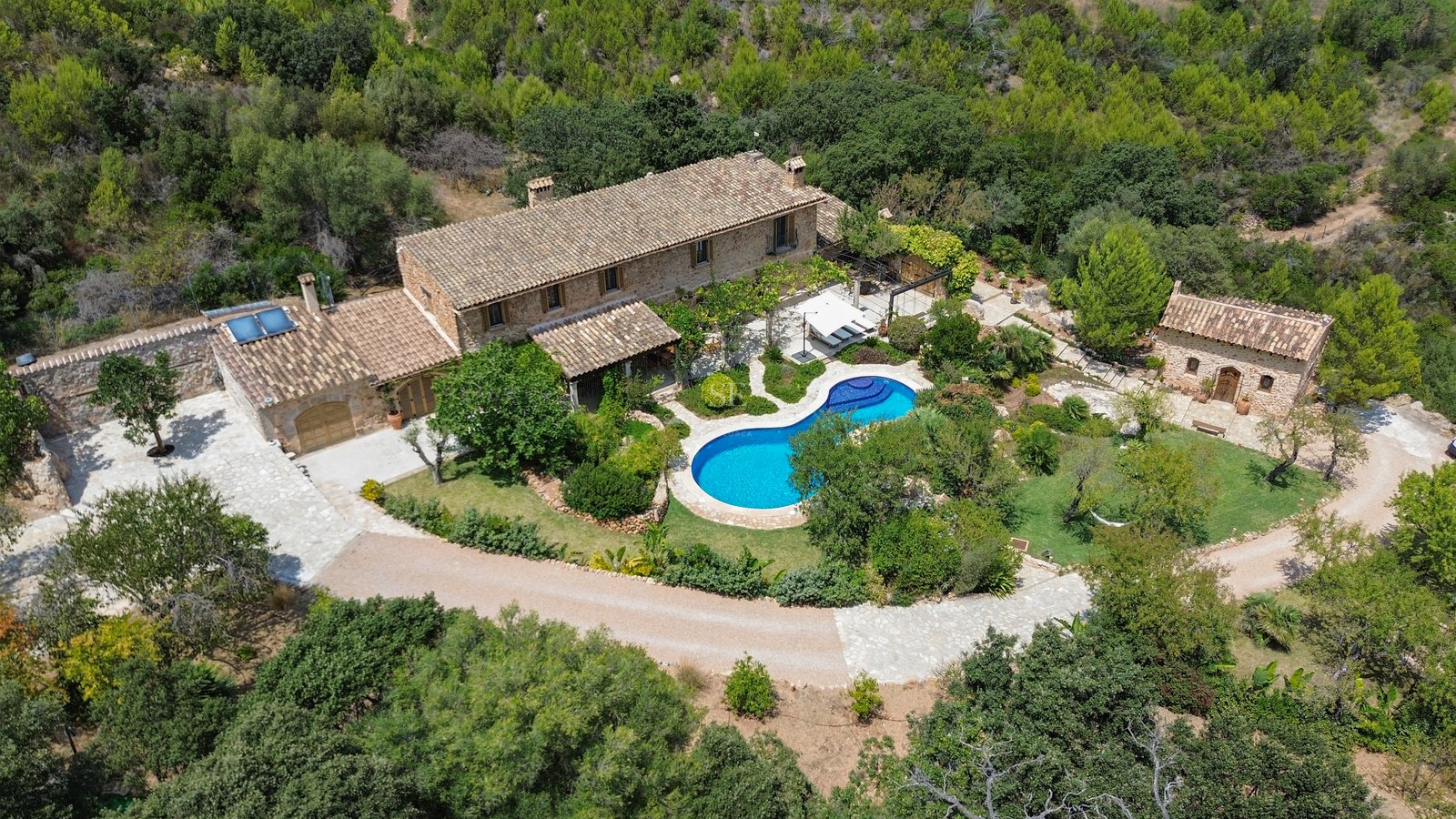 Aerial shot of a stone villa with a blue pool, guest house, and gardens surrounded by green forest.