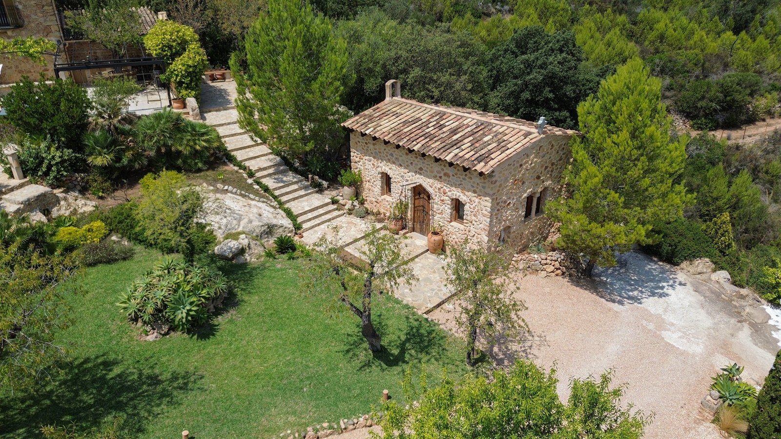 Vista aérea de una casita de piedra rústica con tejado de terracota rodeada de jardines y árboles mediterráneos.