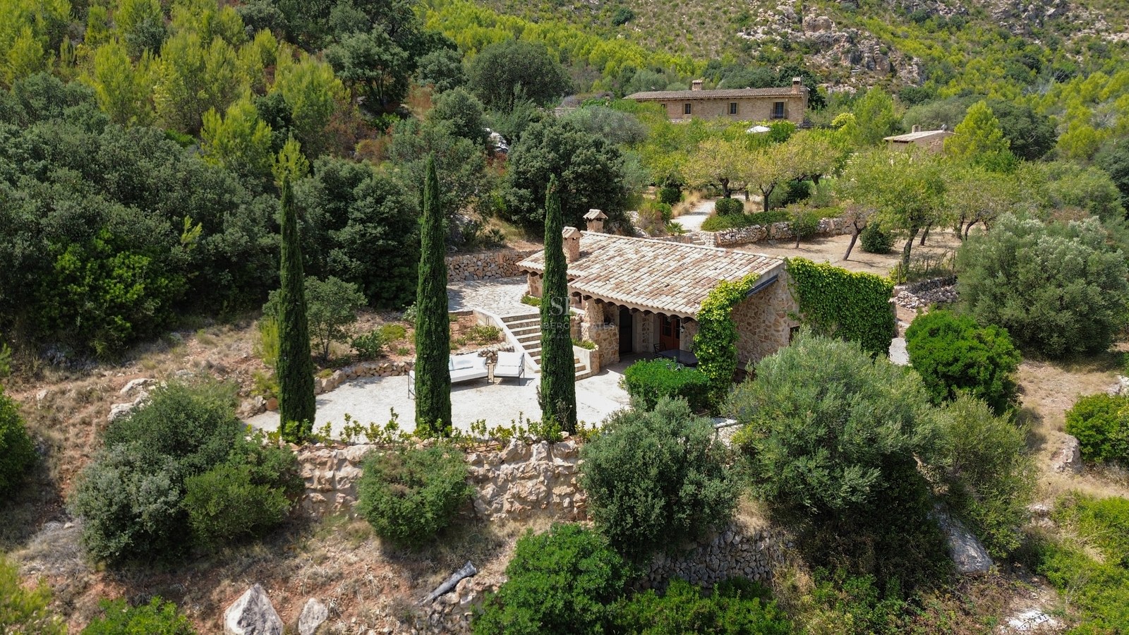 Aerial view of a rustic stone house with a terracotta roof, surrounded by cypress trees and lush green forest.