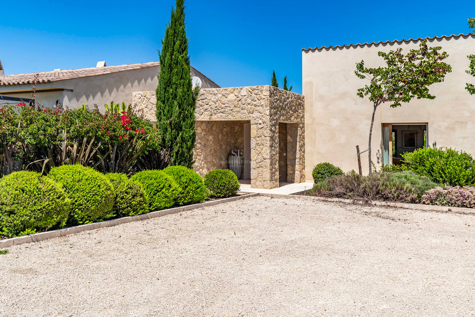 Entrée de villa en pierre avec allée de gravier et arbustes verts sous un ciel bleu.