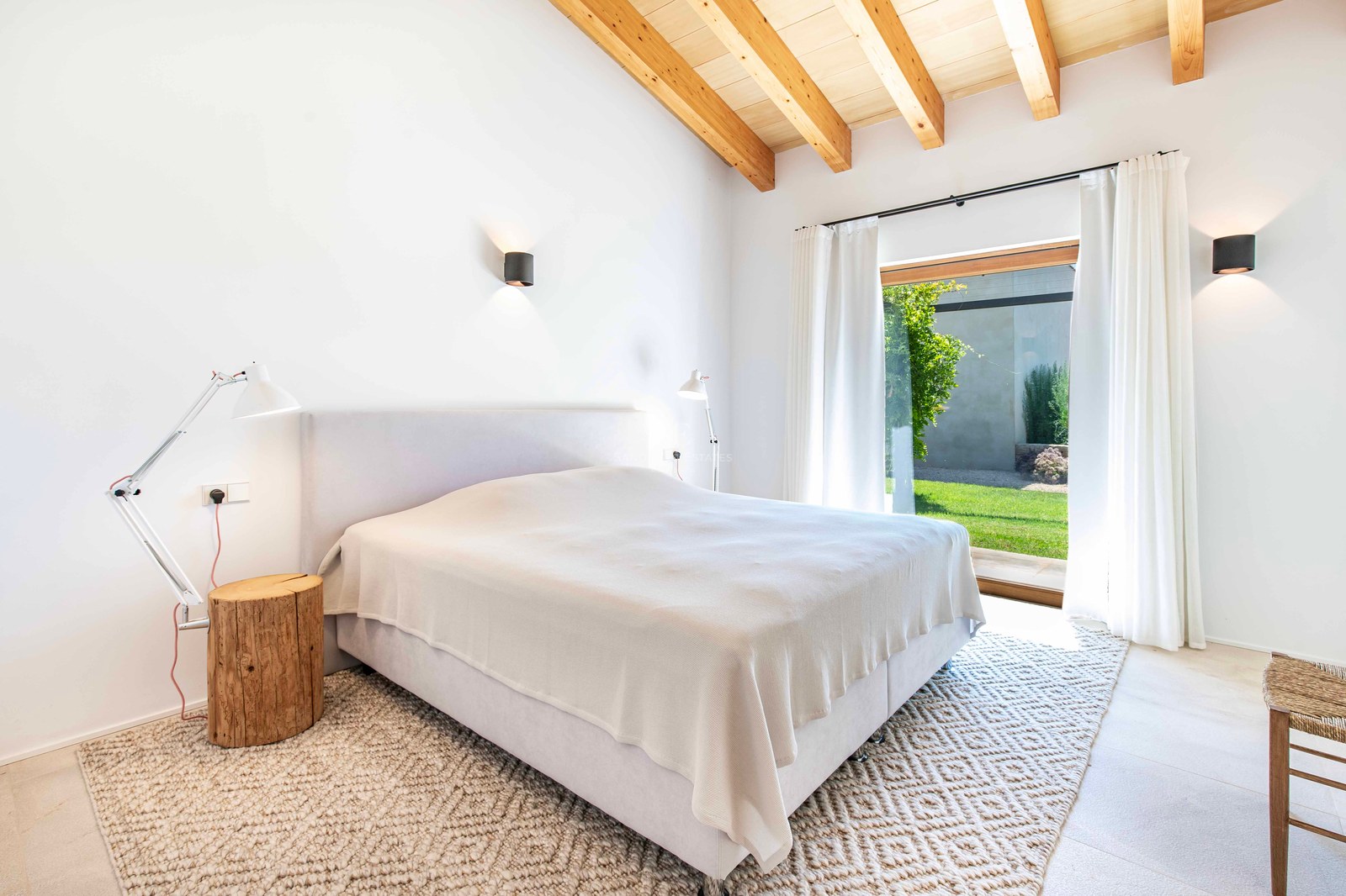 Chambre lumineuse avec plafond voûté en bois, murs blancs, grande baie vitrée donnant sur un jardin et tapis texturé.