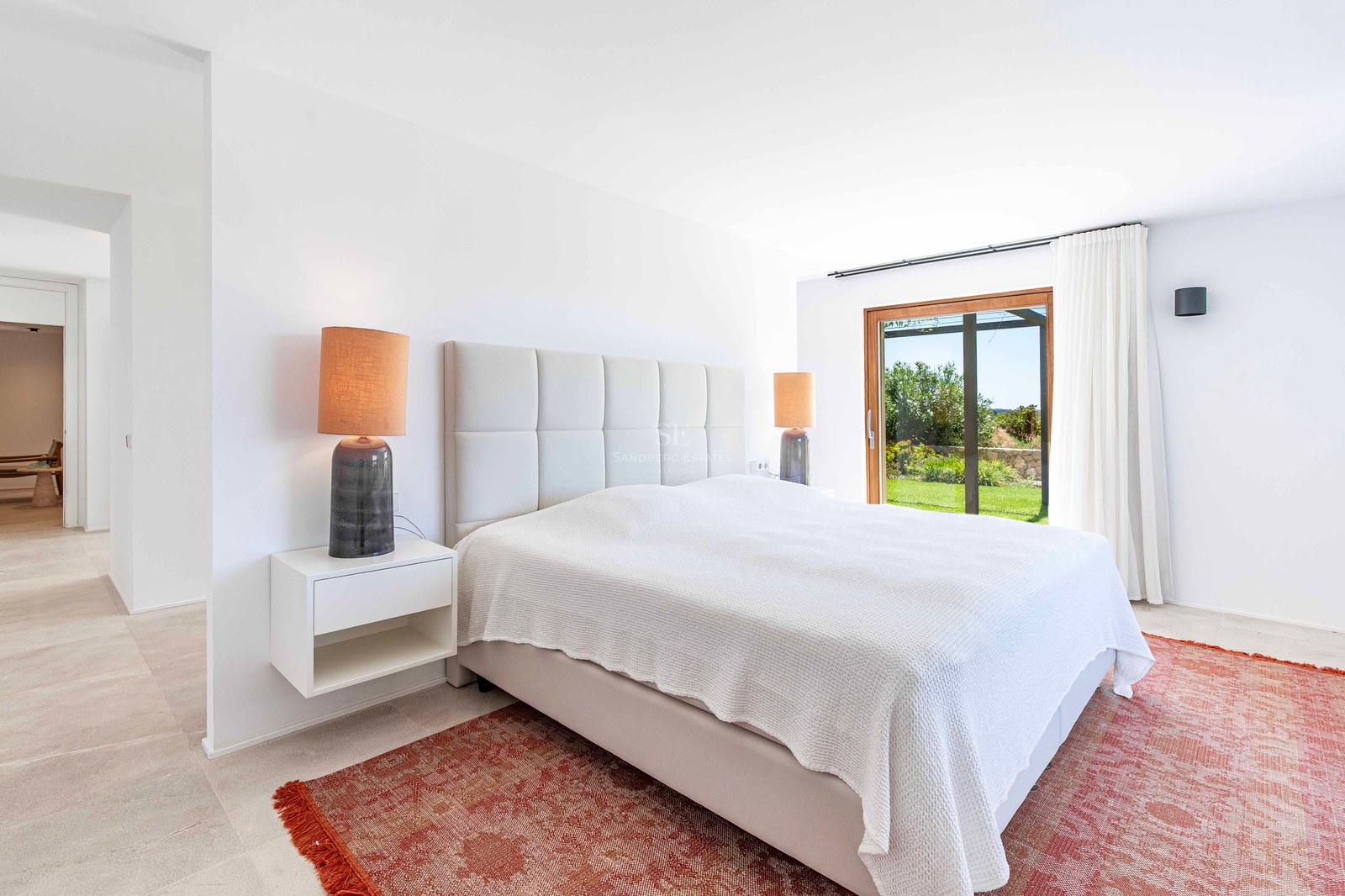 Chambre moderne lumineuse avec grand lit, tapis terre cuite et baies vitrées ouvrant sur un jardin verdoyant.