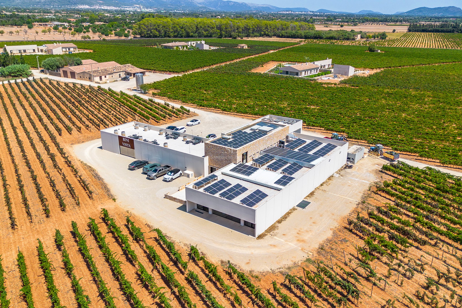 Vue aérienne d'un bâtiment moderne avec panneaux solaires entouré de vignobles verdoyants et de montagnes.