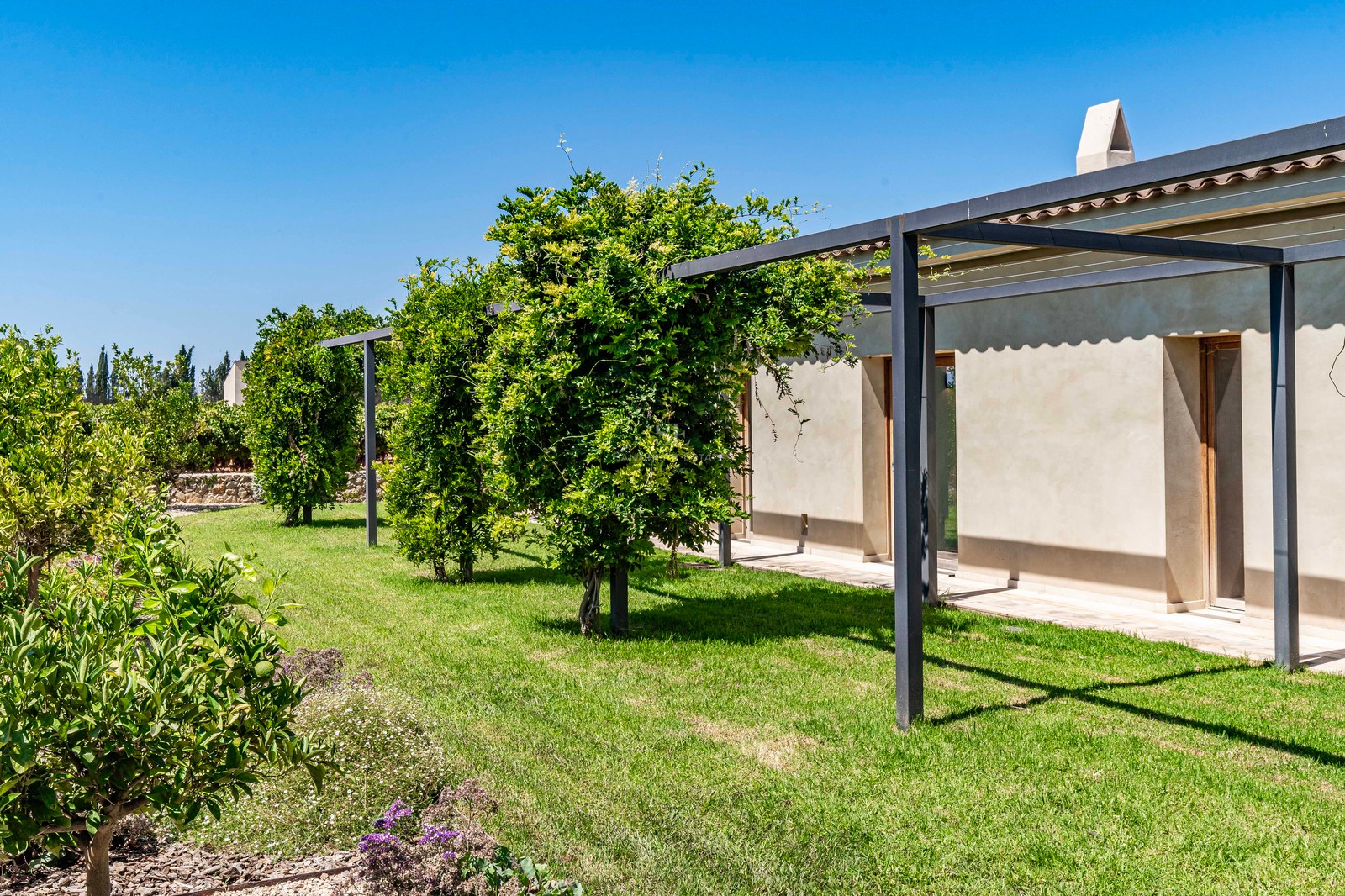 Extérieur d'une villa moderne couleur sable avec pergola en métal noir, pelouse verte et arbres sous un ciel bleu clair.
