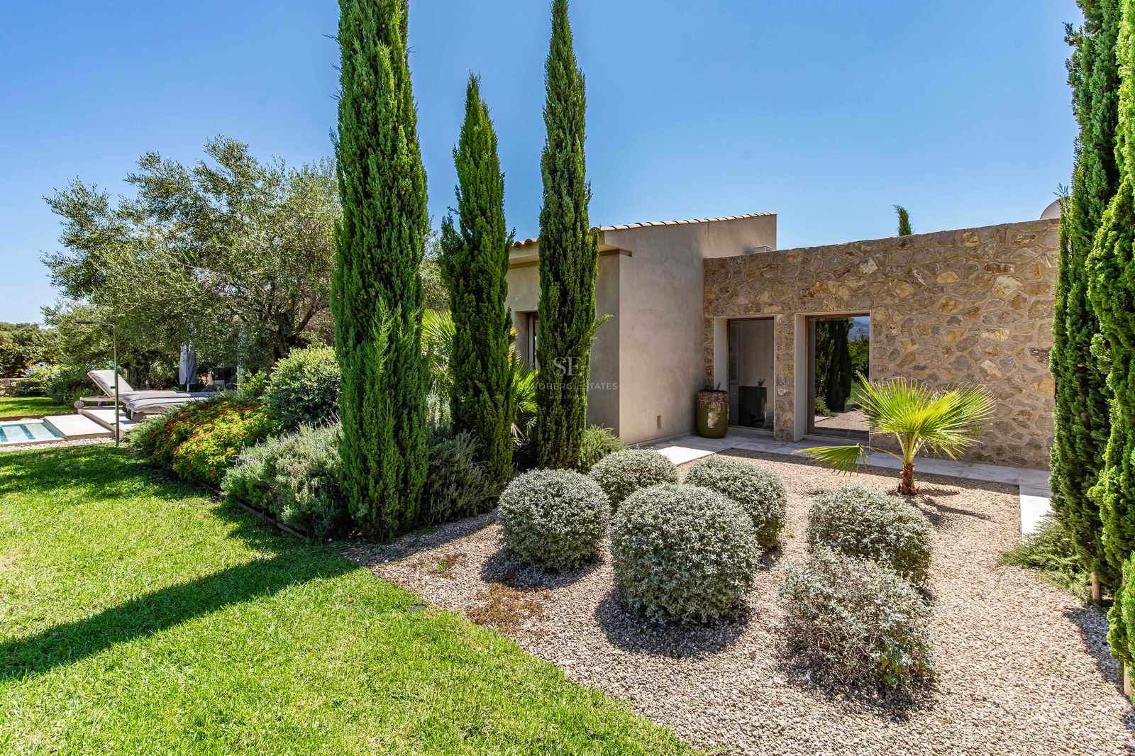 Villa moderne aux murs en pierre naturelle et jardin avec cyprès et arbustes sous un ciel bleu clair.
