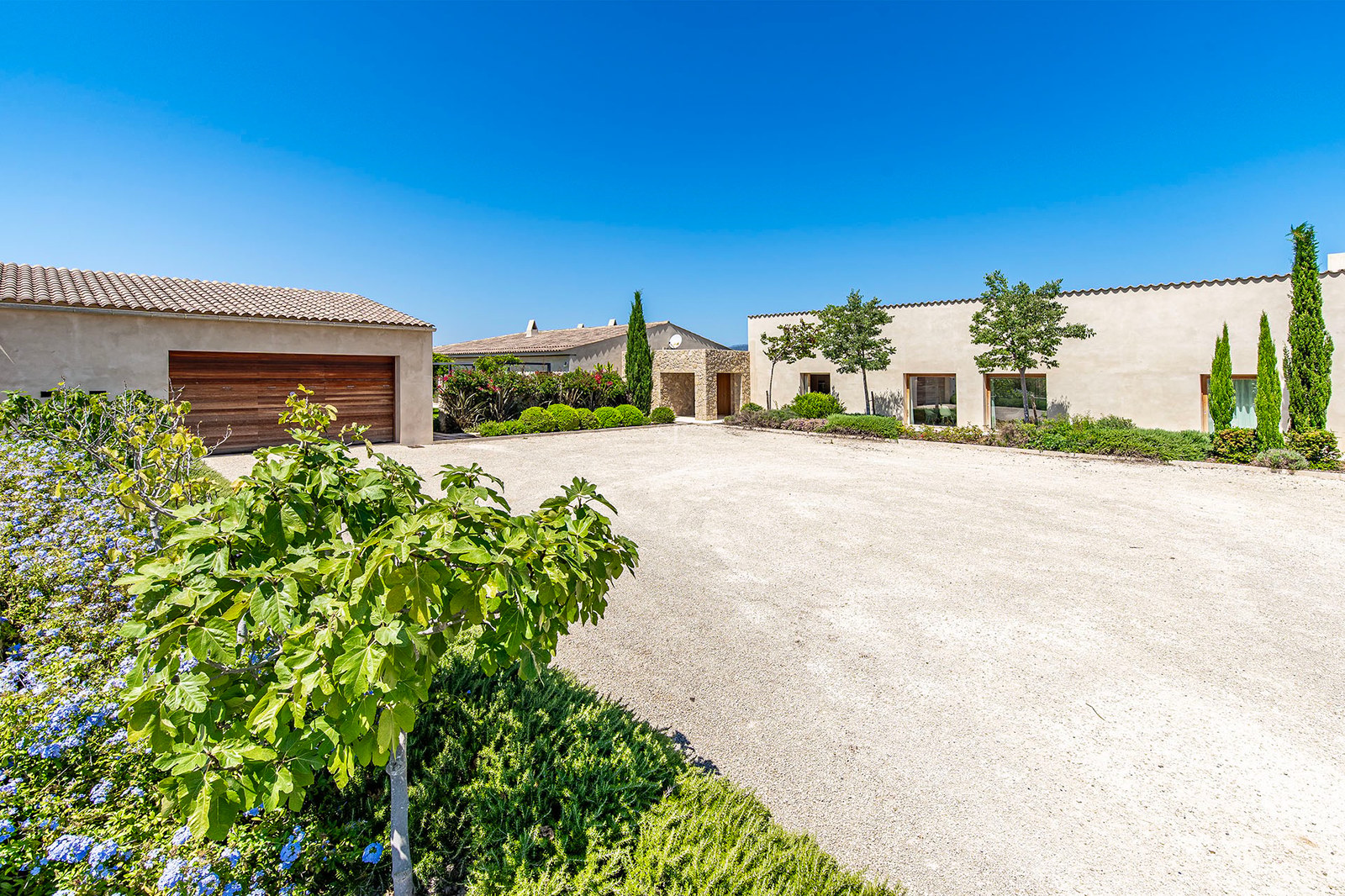 Villa moderne avec façade sable, porte de garage en bois et allée en gravier sous un ciel bleu pur.