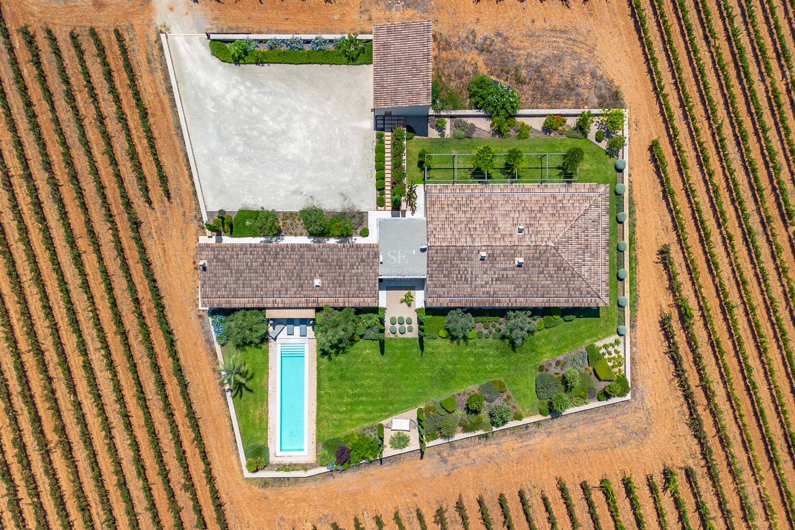 Prise de vue aérienne d'une villa avec piscine turquoise et jardins structurés, entourée de rangées de vignes.