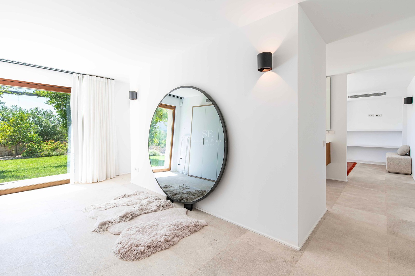 Intérieur minimaliste lumineux avec un grand miroir rond, sol en pierre et baies vitrées ouvrant sur un jardin verdoyant.