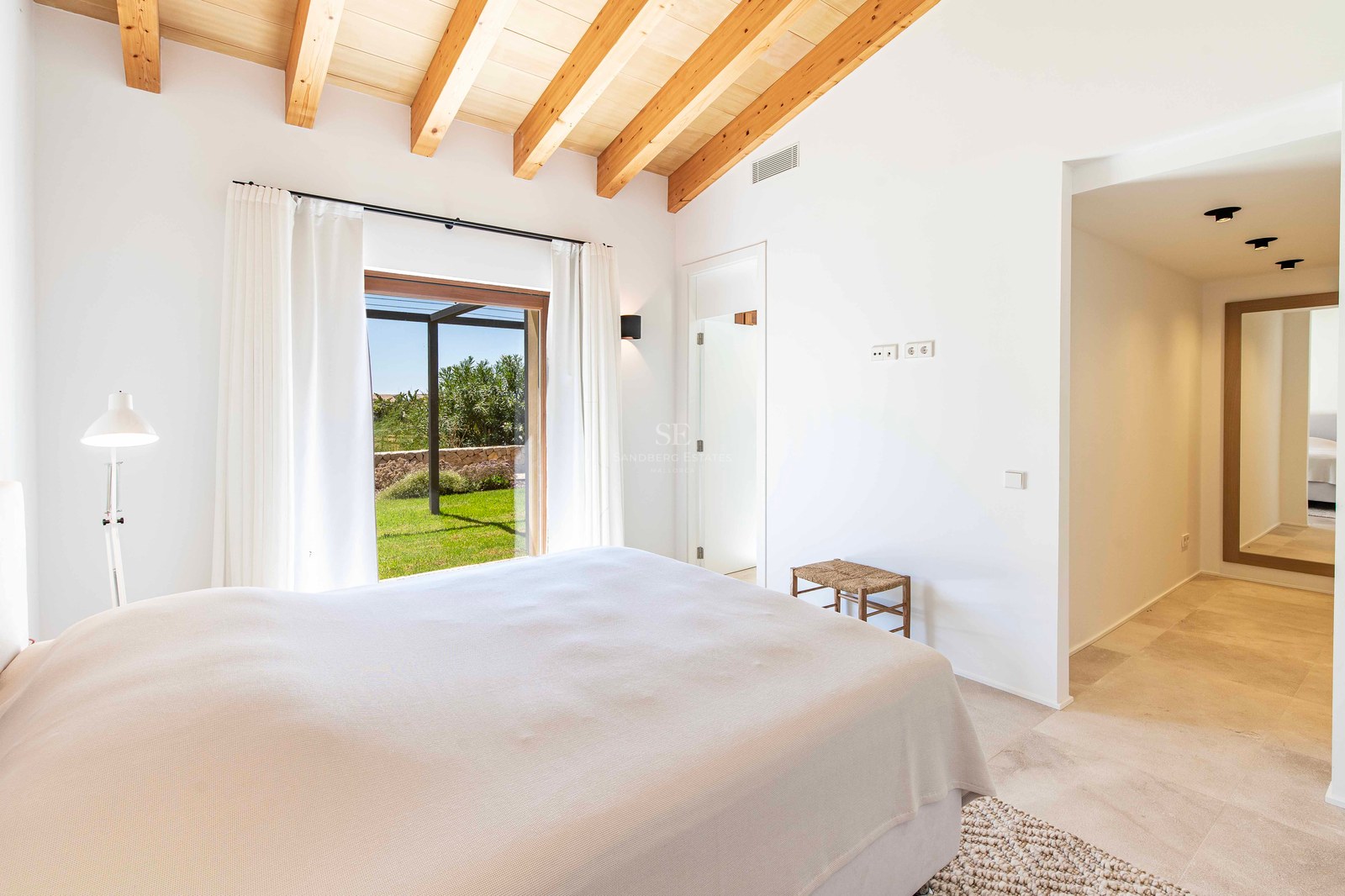 Chambre lumineuse avec poutres en bois, sol en pierre et accès direct à un jardin verdoyant par une porte vitrée.