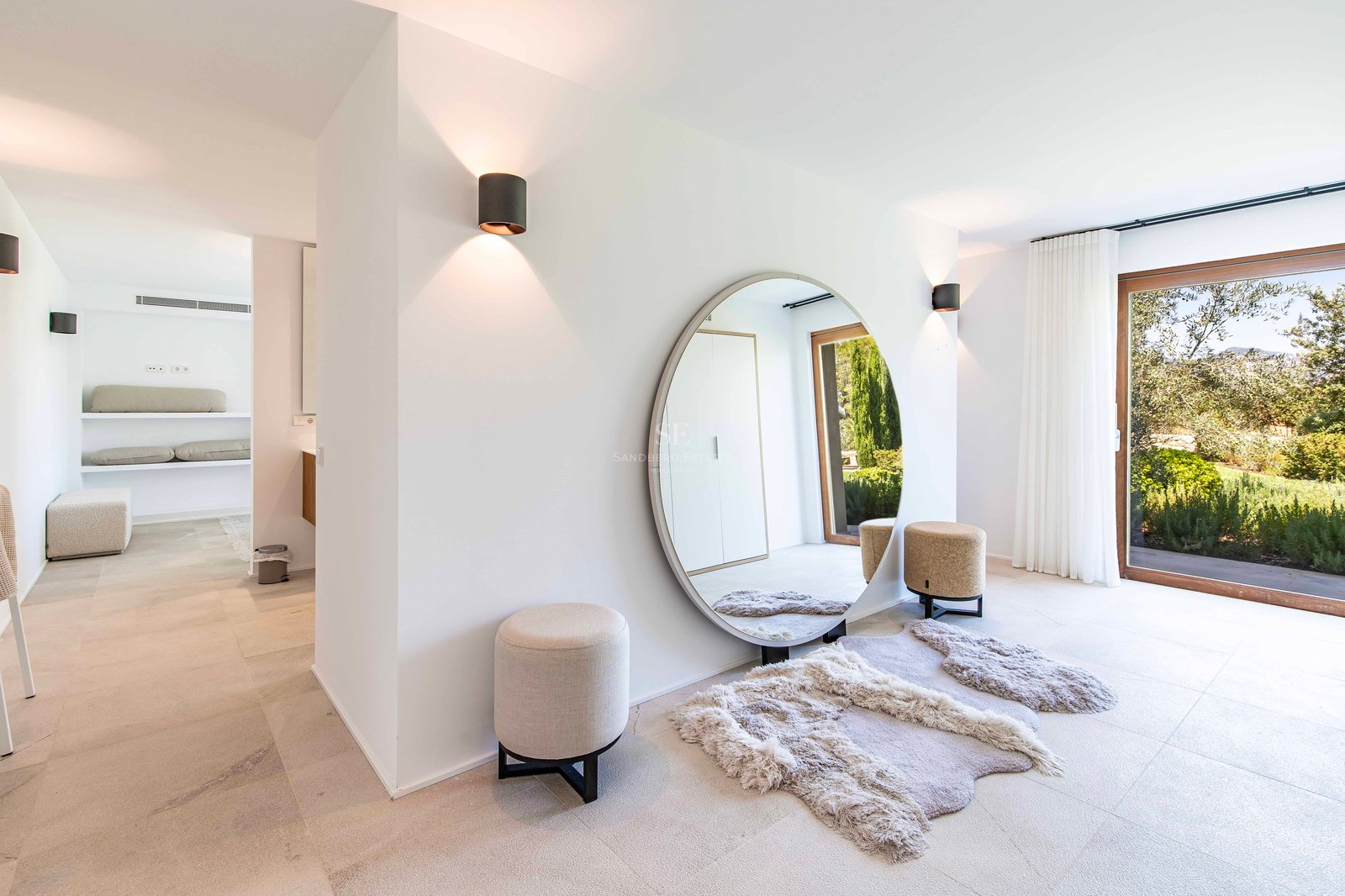 Couloir minimaliste avec grand miroir circulaire, poufs crème et vue sur un jardin via une porte vitrée.