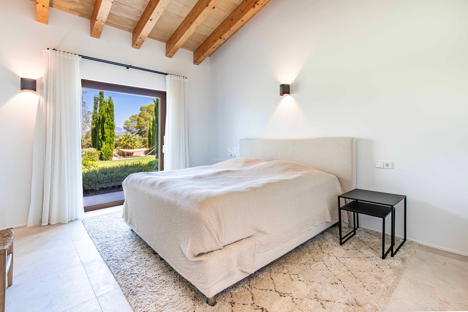 Chambre moderne avec poutres apparentes, lit blanc et grande baie vitrée ouvrant sur un jardin luxuriant.