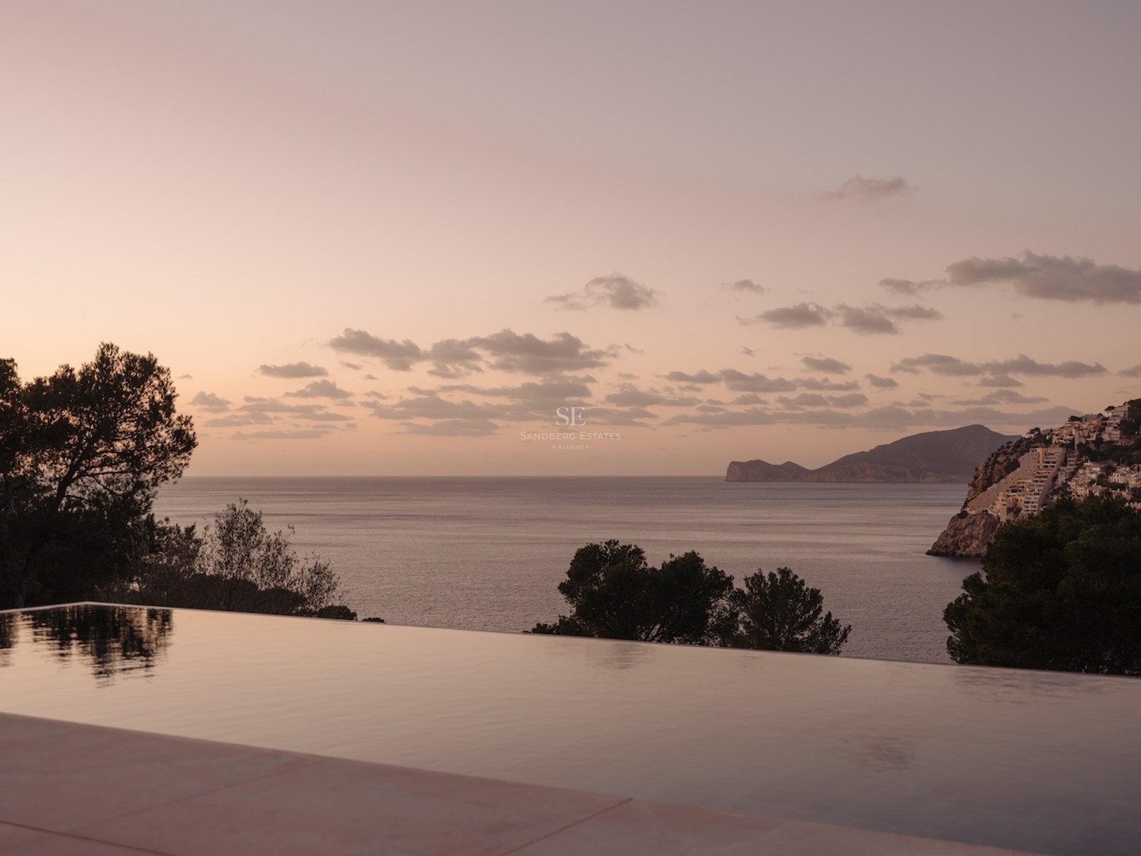 Piscina infinita reflejando un cielo rosado al atardecer con vistas a la costa y el mar Mediterráneo.