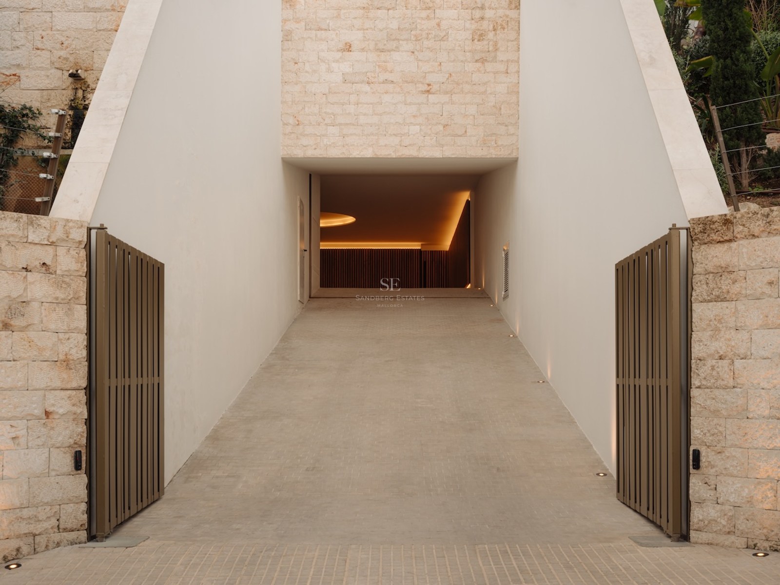 Entrada de garaje moderna con muros de piedra natural y portones metálicos que conducen a un parking iluminado.