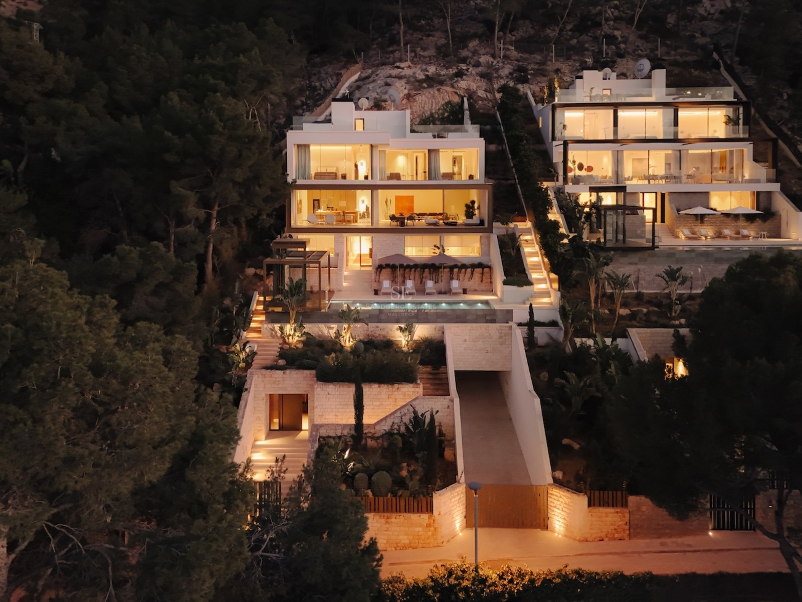 Villa moderna de varios niveles con fachada iluminada, piscina y terrazas en una ladera boscosa de noche.