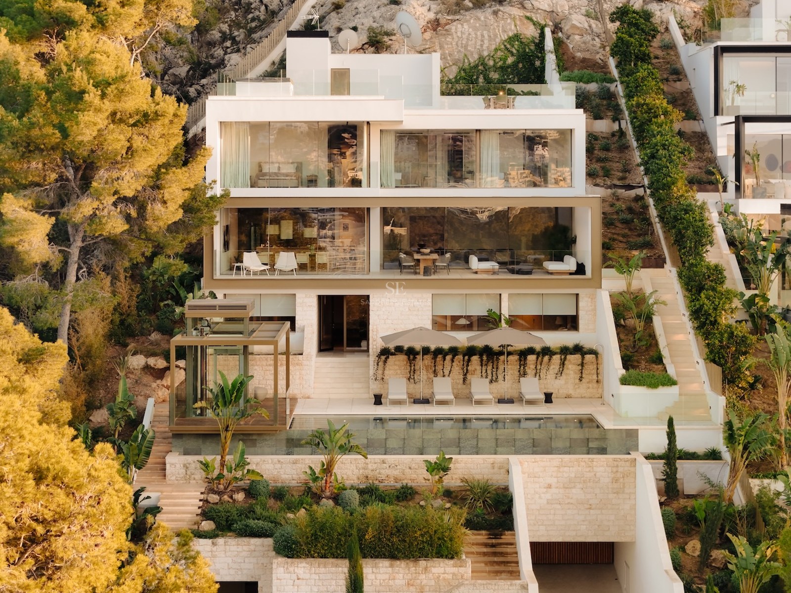 Fachada exterior de una villa moderna de cuatro niveles con muros de piedra natural, ventanales y piscina infinita.