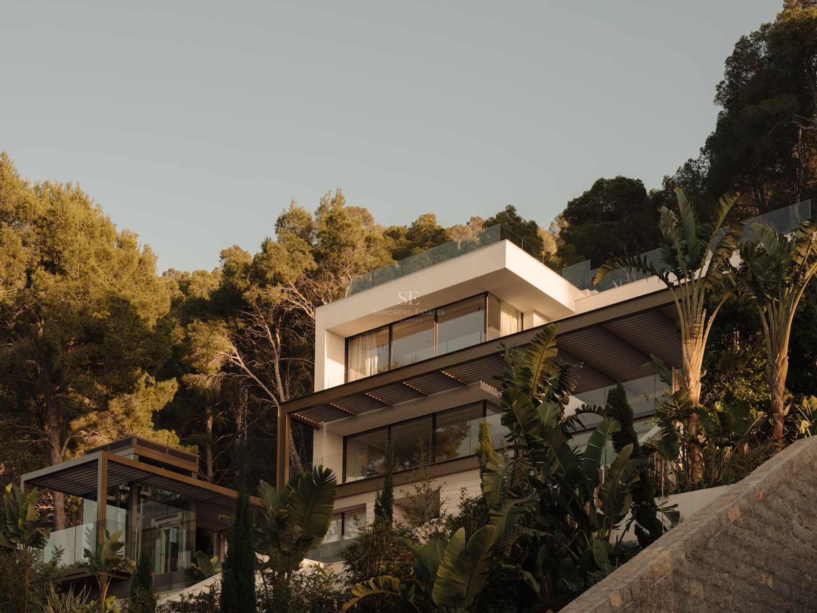 Villa blanca moderna de varios niveles con barandillas de cristal rodeada de pinos y plantas tropicales.