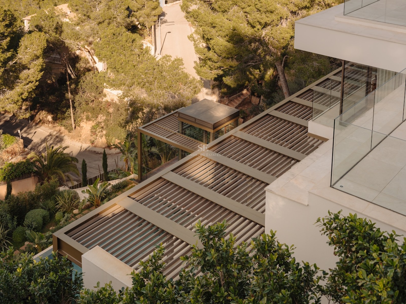 Vista elevada de una villa moderna con pérgola de lamas, barandillas de cristal y entorno boscoso.