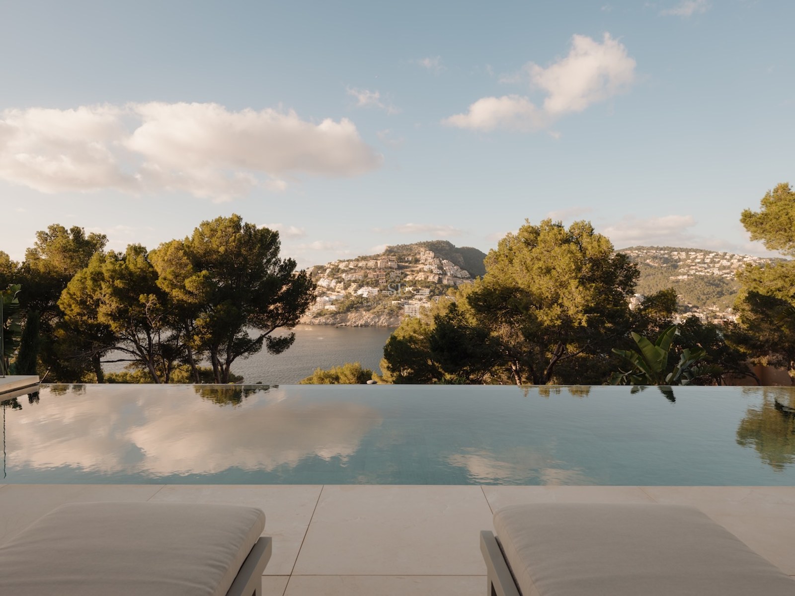 Piscina infinita con vistas a una bahía mediterránea y colinas con pinos, hamacas en primer plano.