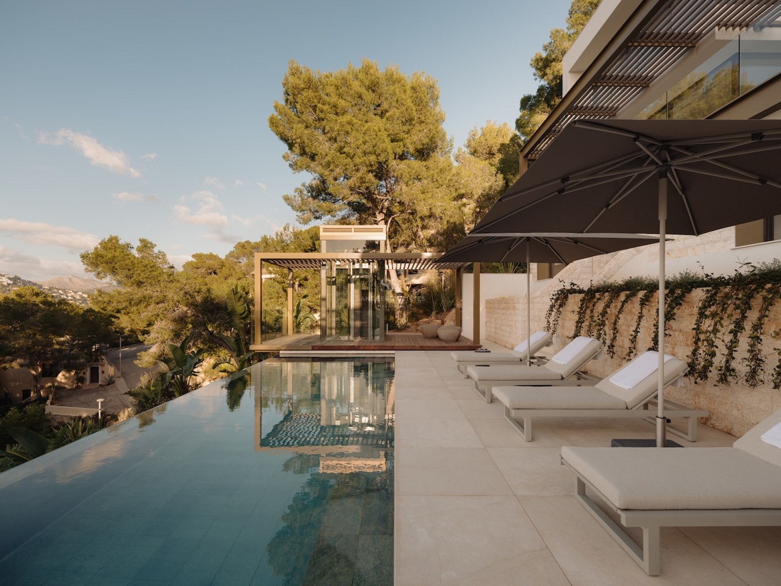 Piscina infinita con cuatro tumbonas y sombrillas en una terraza de piedra natural rodeada de pinos mediterráneos.