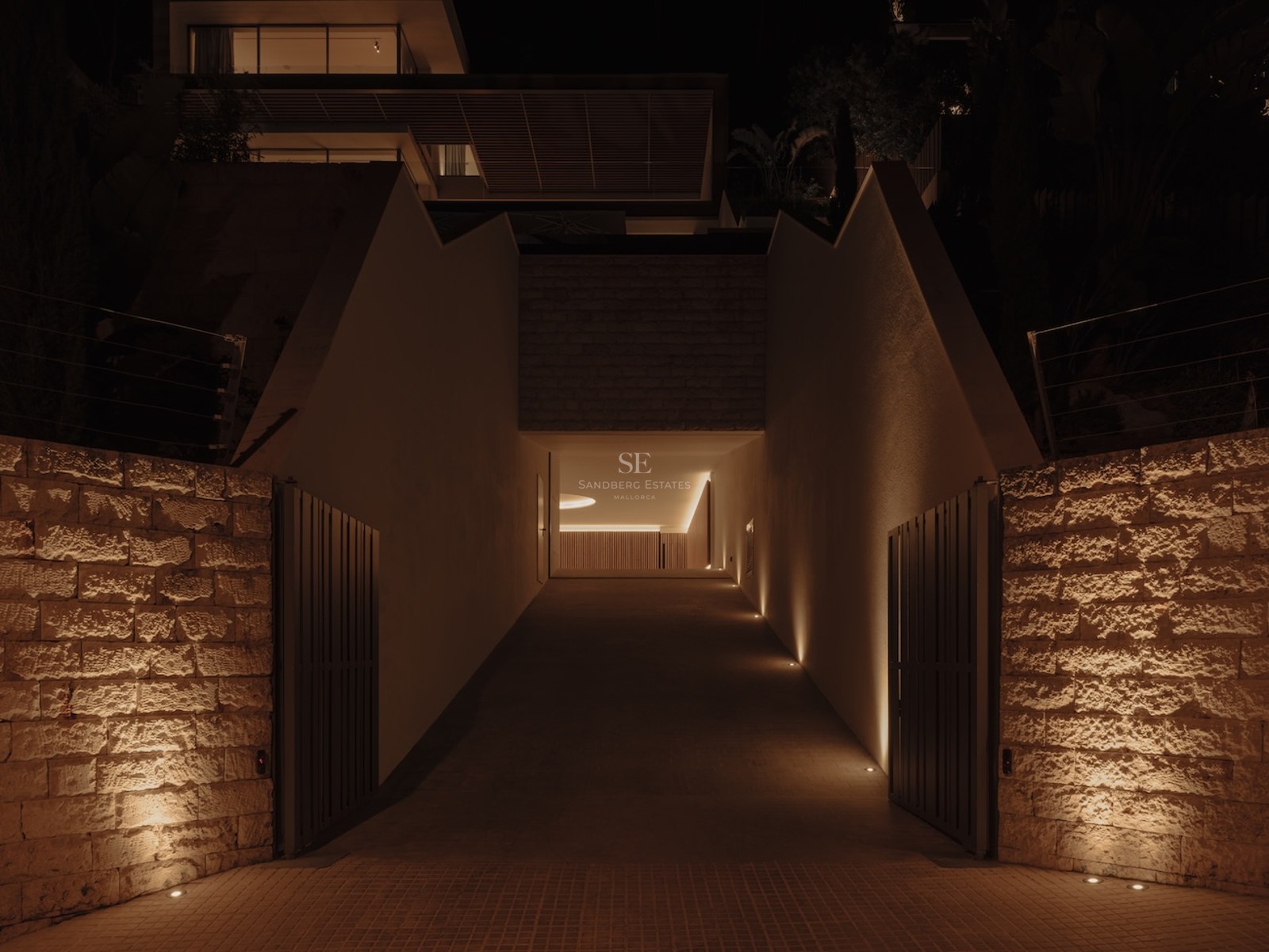 Entrada nocturna de una villa moderna con luces empotradas en paredes de piedra natural y puertas de metal oscuro.