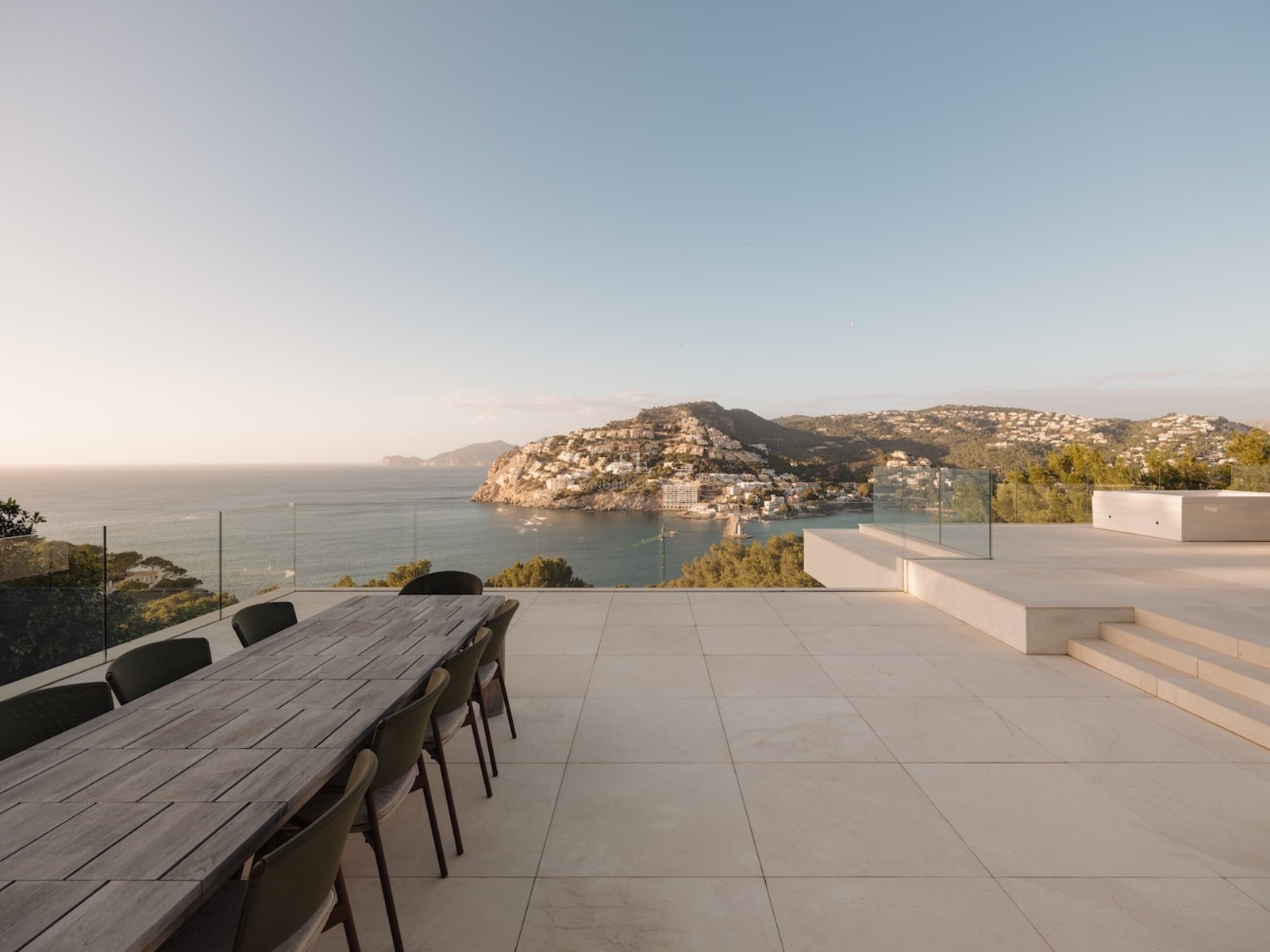 Terraza de piedra elevada con mesa de comedor de madera con vistas a una bahía mediterránea y montañas al atardecer.