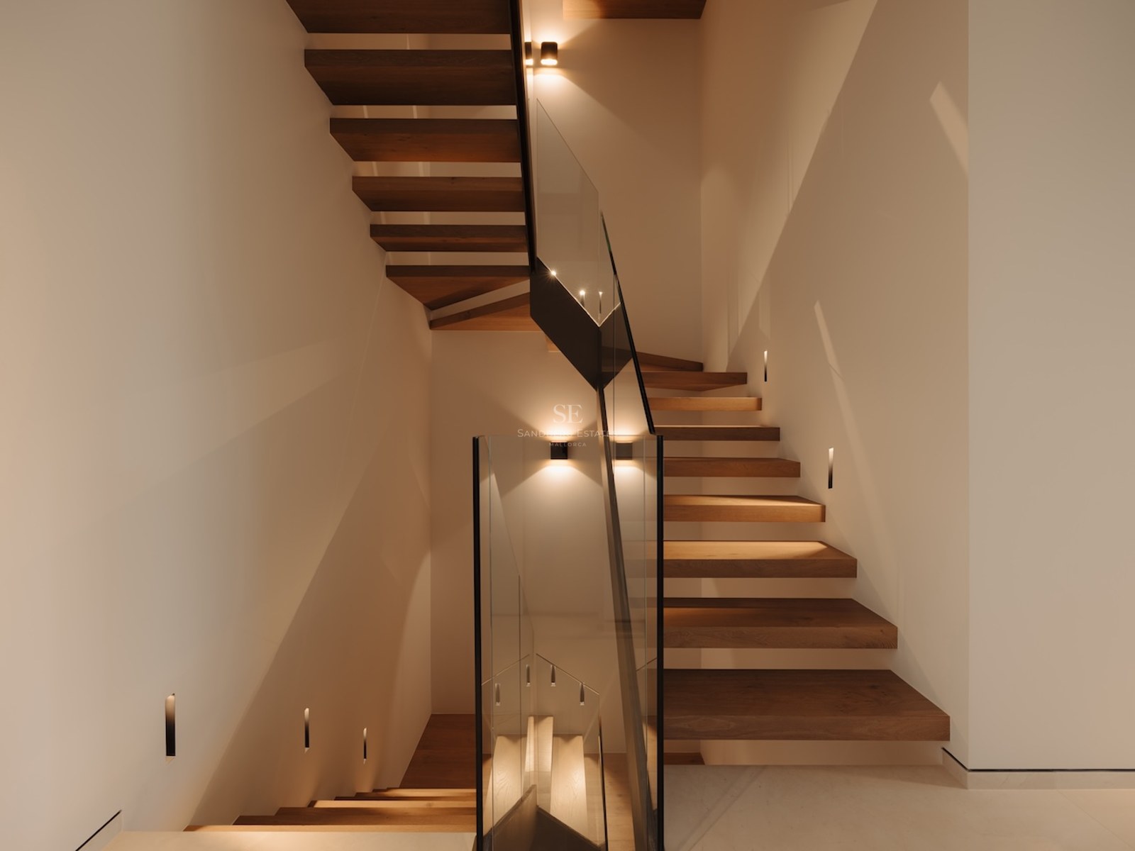 Escalera moderna con peldaños de roble y barandillas de cristal, iluminada con luces LED empotradas.