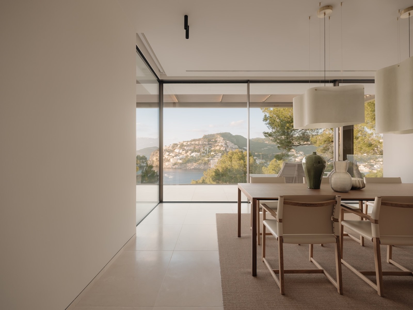 Comedor moderno con mesa de madera, sillas de cuero y grandes ventanales con vistas a una bahía mediterránea.