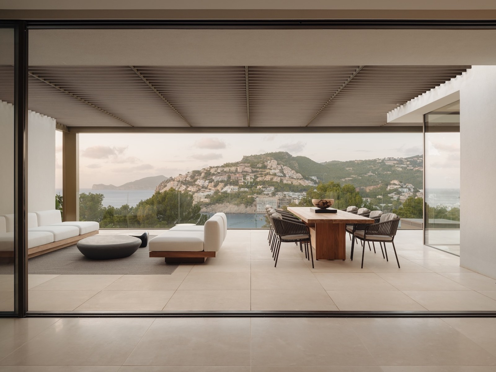 Terraza moderna cubierta con mesa de comedor de madera, sofás blancos y vistas al mar tras barandillas de cristal.