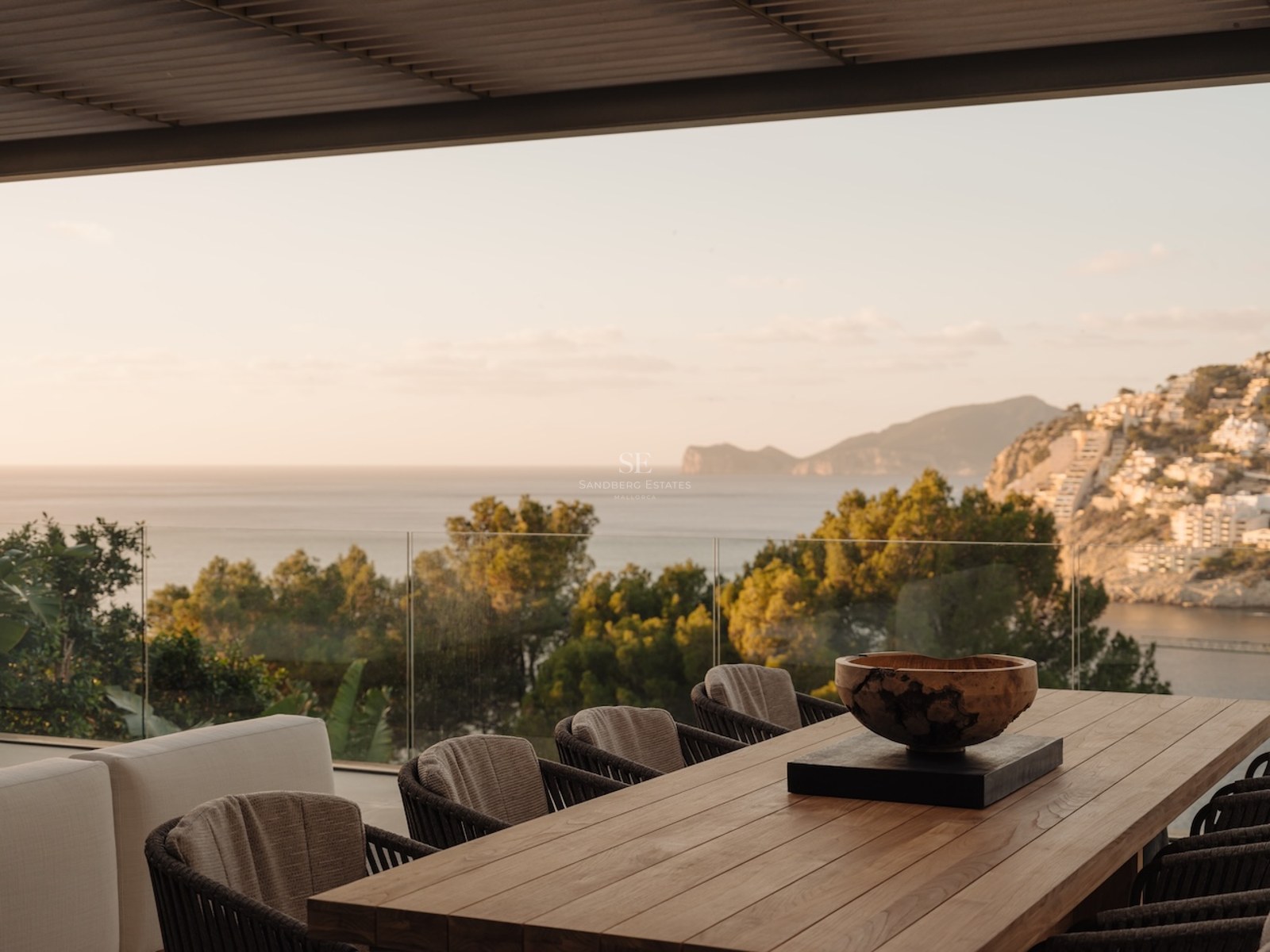 Terraza cubierta con gran mesa de comedor de madera y sillas contemporáneas con vistas al mar Mediterráneo y la costa.