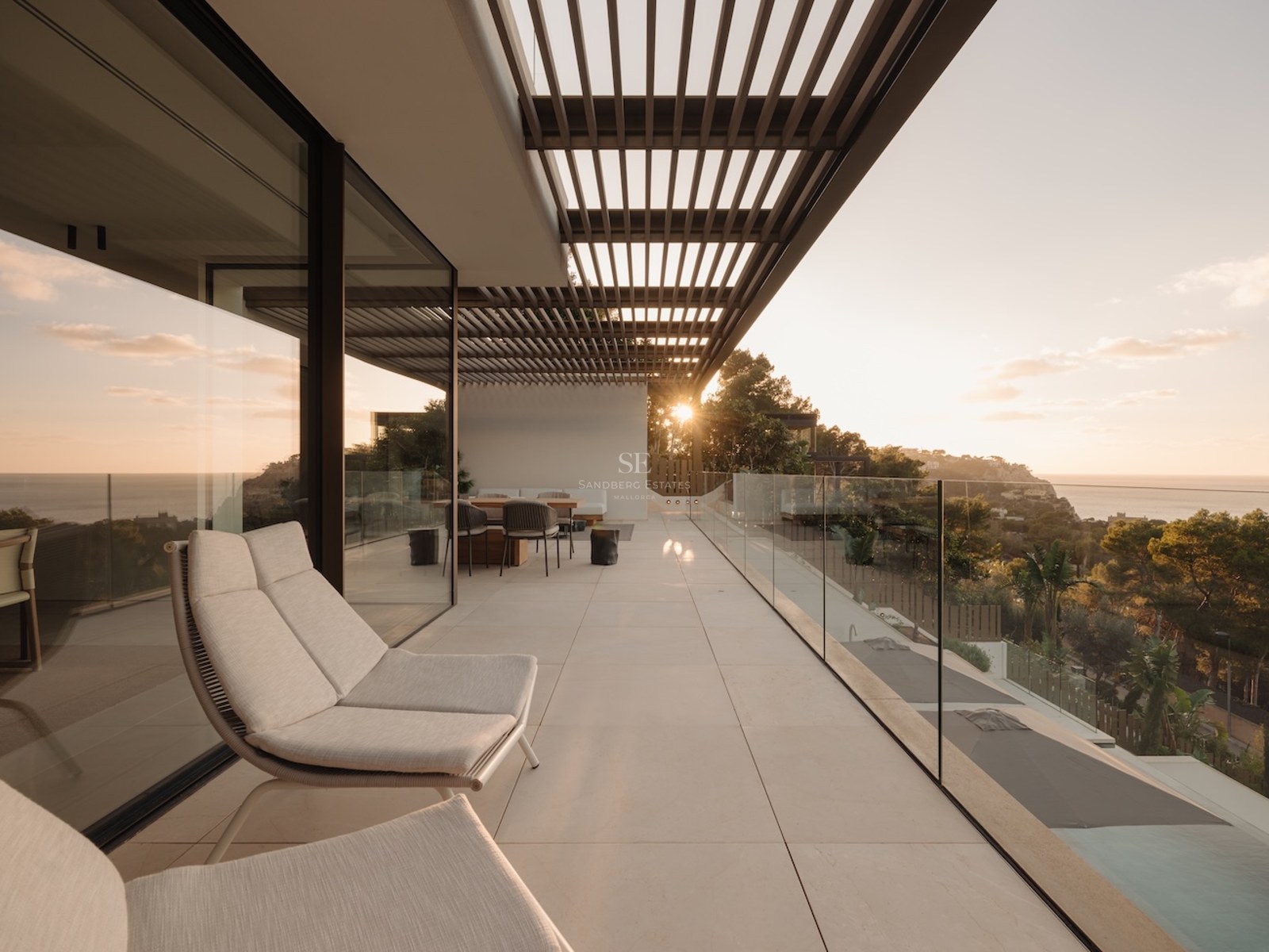 Terraza contemporánea con barandillas de cristal, tumbonas y pérgola con vistas al mar durante el atardecer.