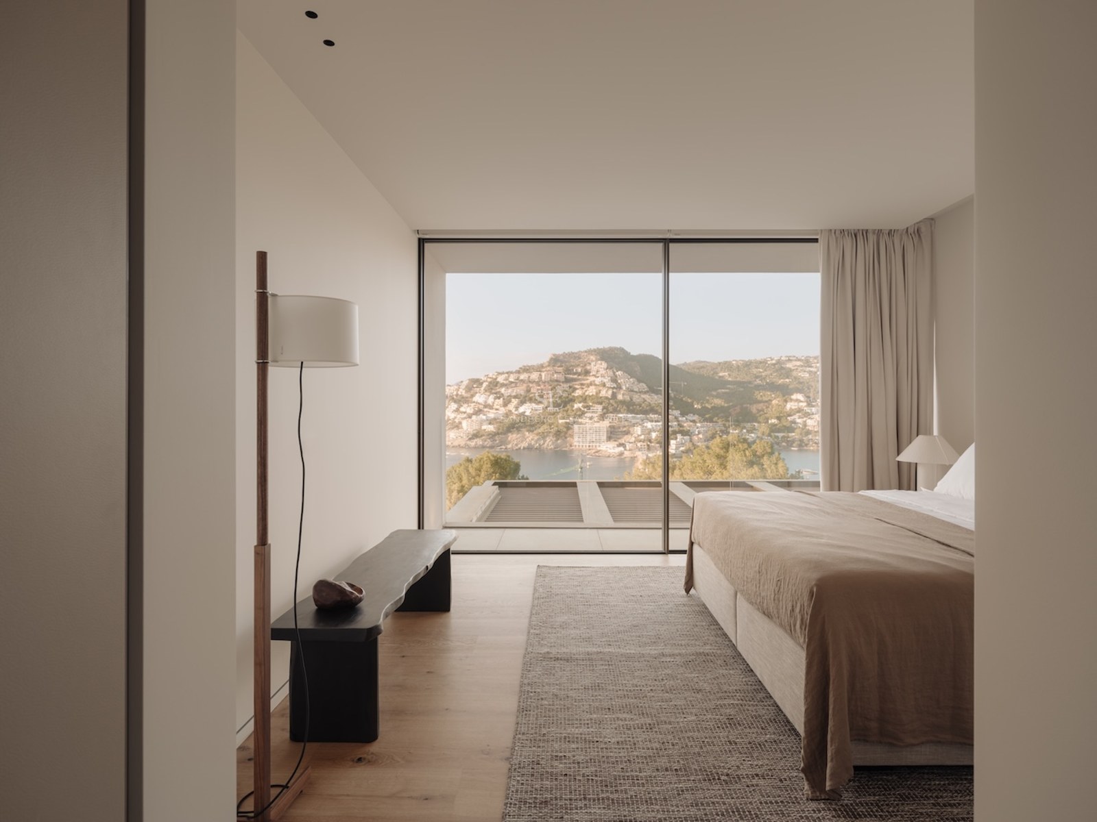 Dormitorio moderno con ventanales de suelo a techo frente a una bahía mediterránea, tonos neutros y suelos de roble.