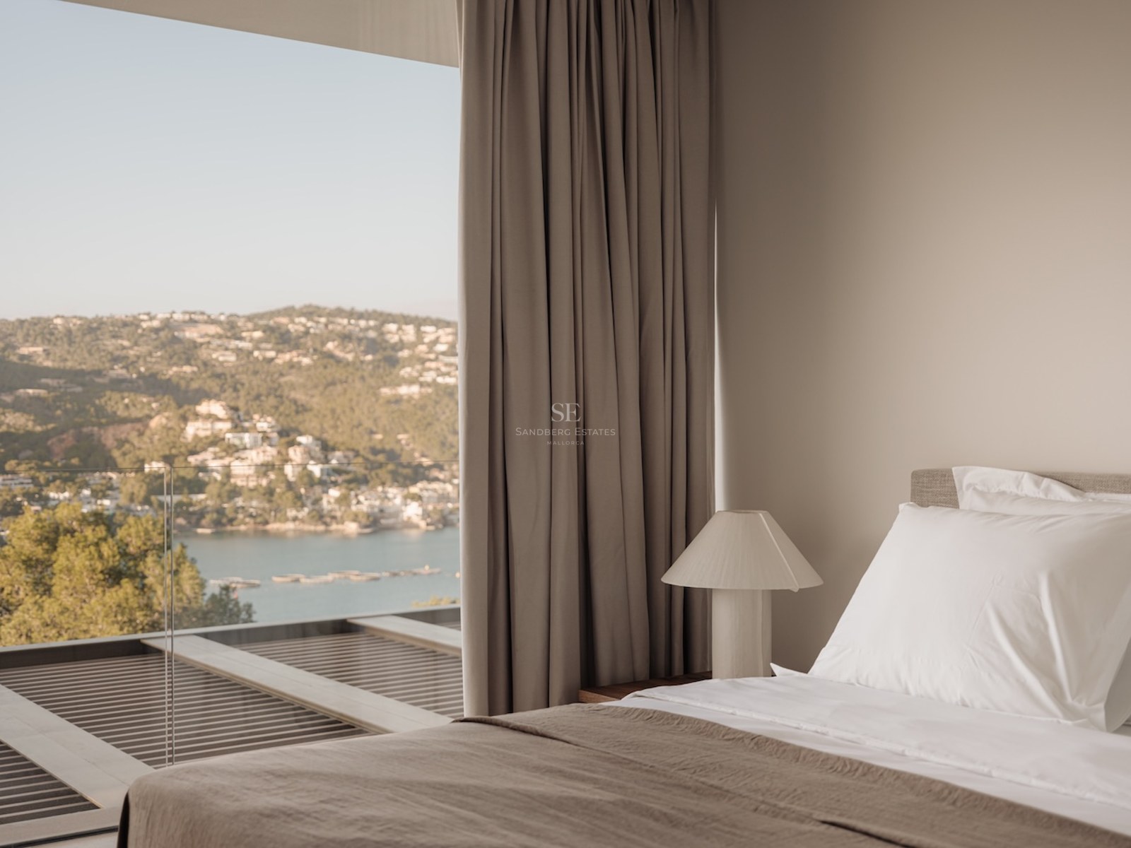 Dormitorio moderno con sábanas blancas y ventanales de suelo a techo con vistas a una bahía mediterránea.