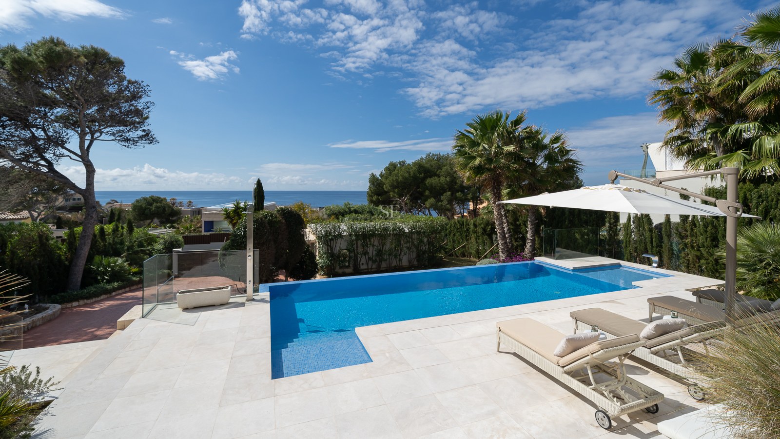 Blue outdoor swimming pool with sun loungers and light stone terrace overlooking the sea and palm trees.