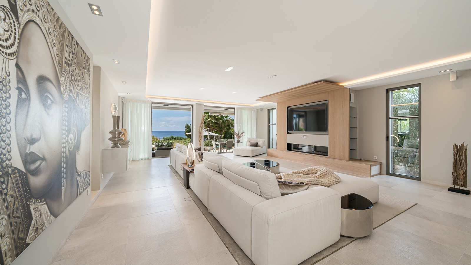 Modern living room with white sectional sofa, large wall mural, and glass doors opening to a sea-view terrace.