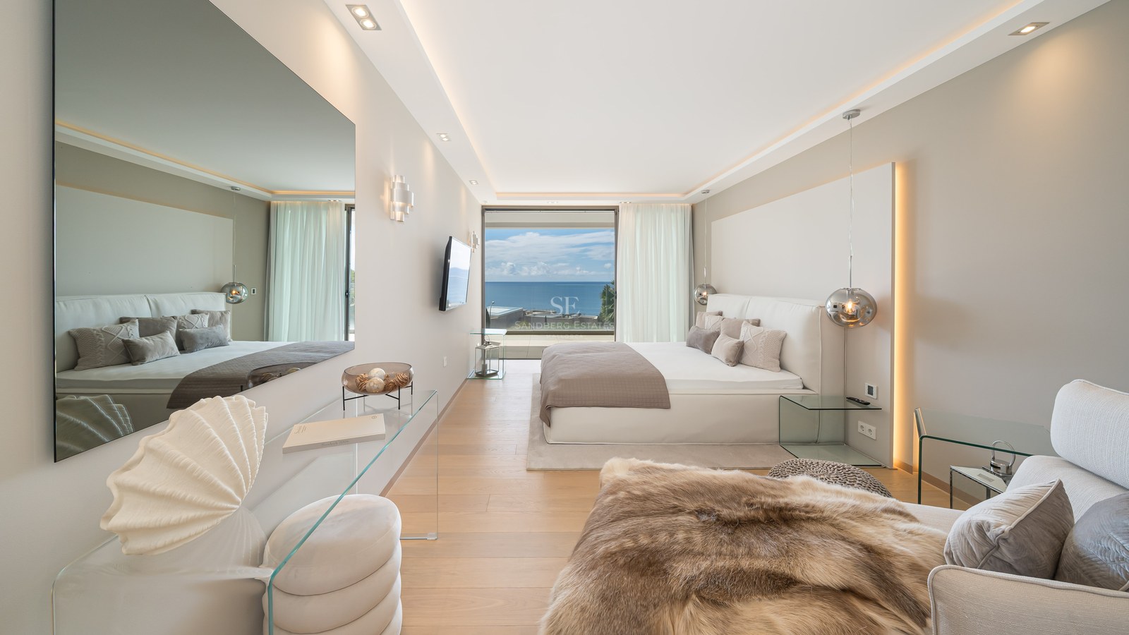 Luxury bedroom with white bed, floor-to-ceiling glass doors overlooking the ocean, glass furniture, and light oak floors.
