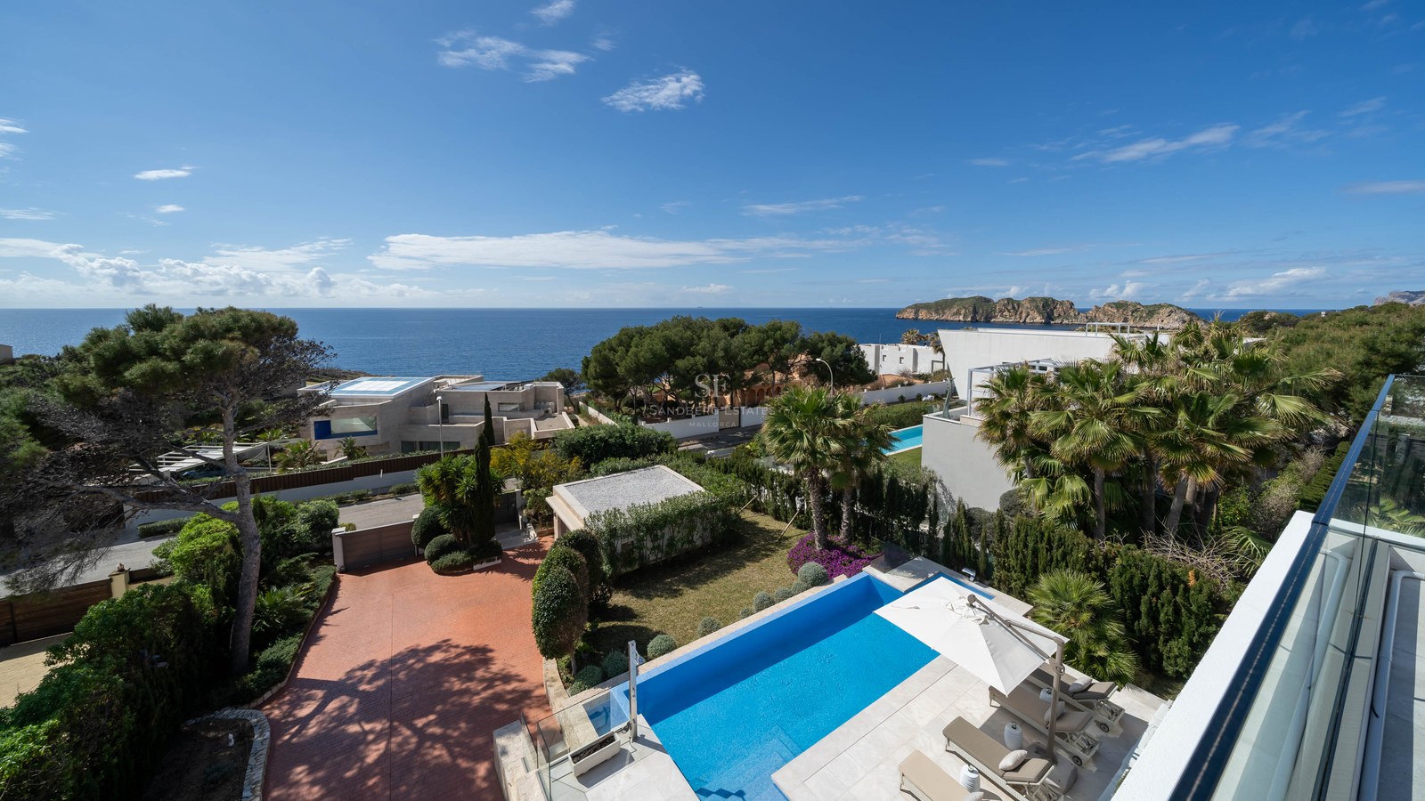 Elevated view of a blue swimming pool, sun deck with loungers, Mediterranean gardens, and sea views under a clear sky.