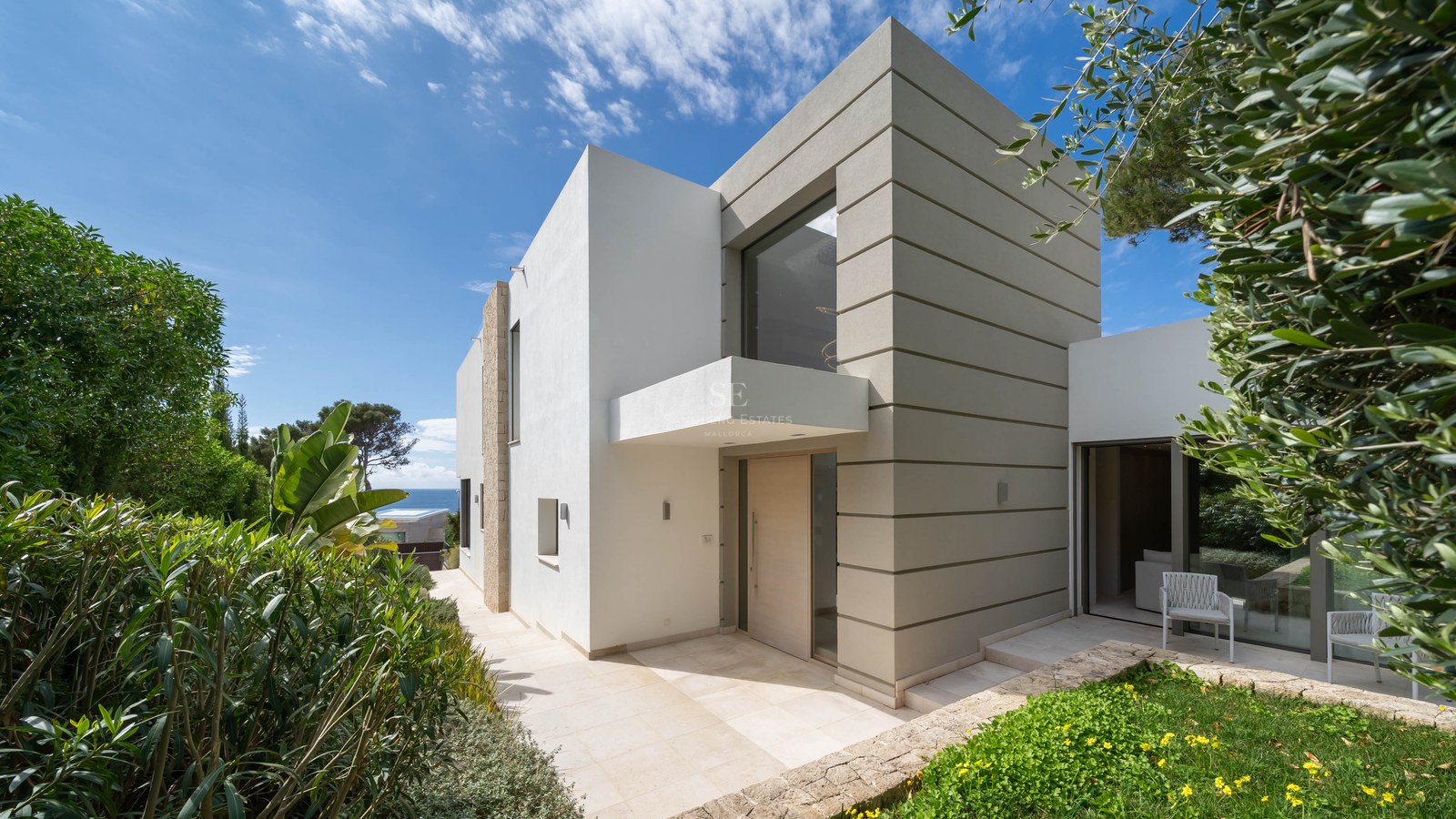 Modern two-story villa facade with white and beige geometric walls, a glass entrance, and Mediterranean landscaping.