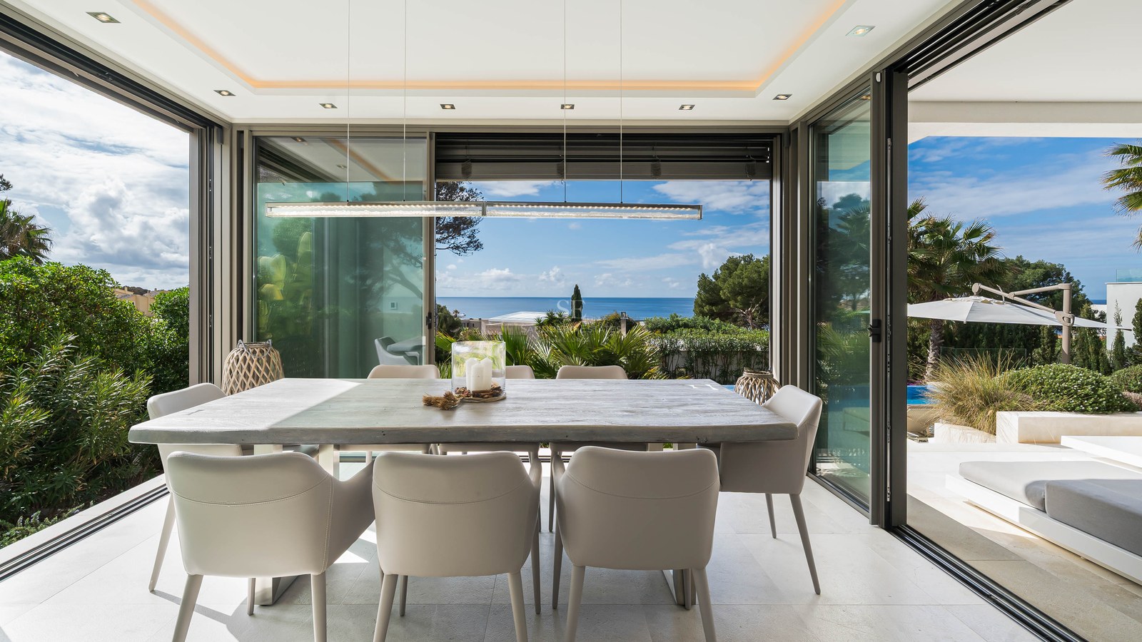 Modern dining room featuring a large wooden table and grey chairs, surrounded by glass walls overlooking the sea.