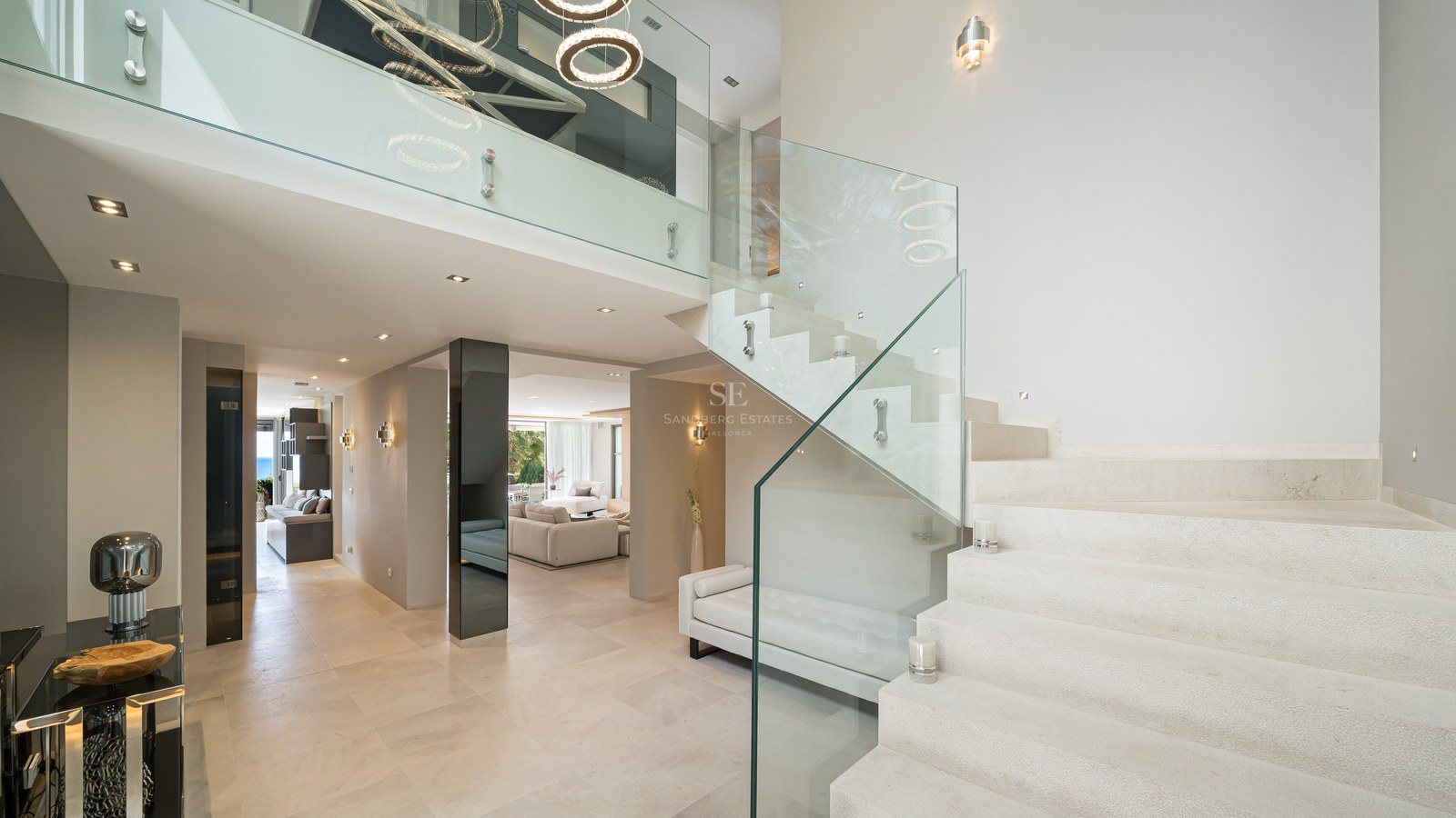 A wide-angle shot of a modern villa entrance featuring a stone staircase with glass balustrades and high ceilings.